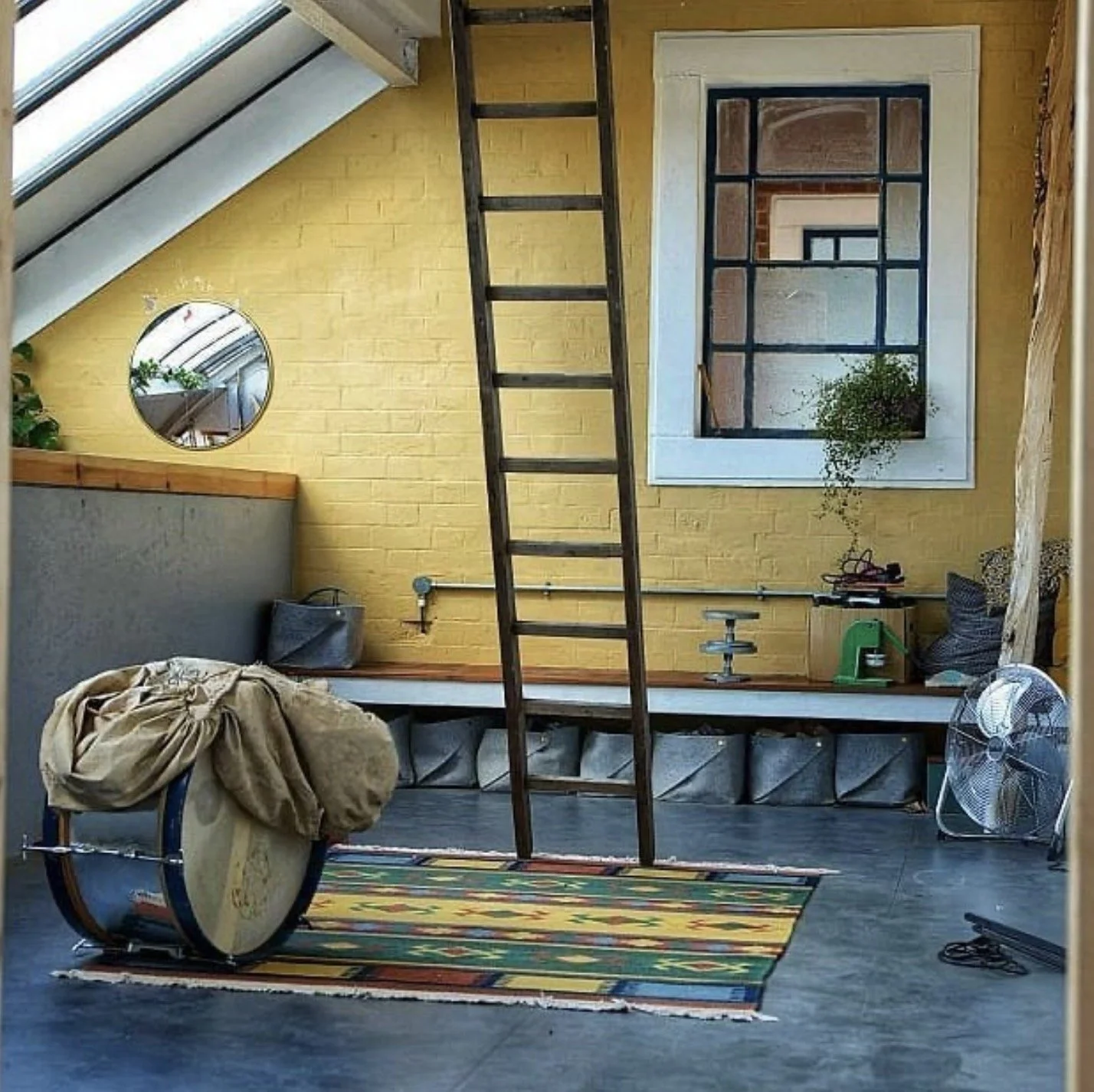 Interior of a room with a sloped ceiling, yellow brick wall, and large window. Contains a drum, a colorful rug, a ladder, a potted plant, and various miscellaneous items.