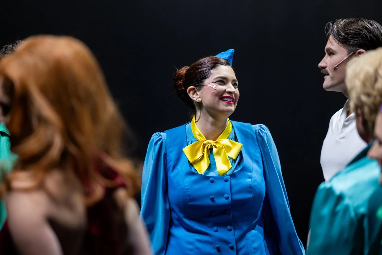 A woman dressed as a colorful clown or performer, smiling and talking to a man, surrounded by other people in costumes, against a black background.
