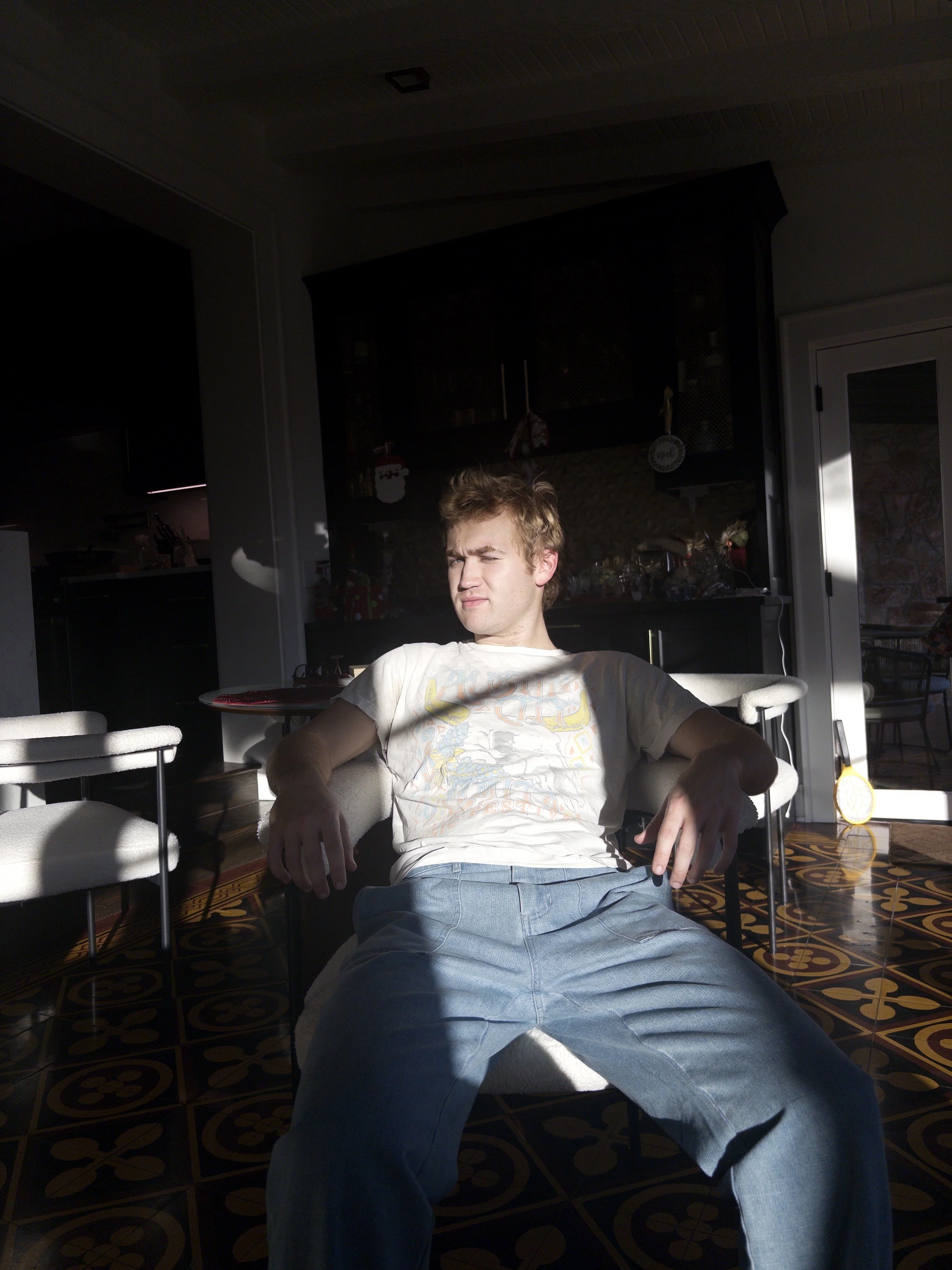 A young man with messy blond hair sitting on a chair in a dimly lit room, with sunlight shining on his face, surrounded by white chairs and dark furniture in the background.