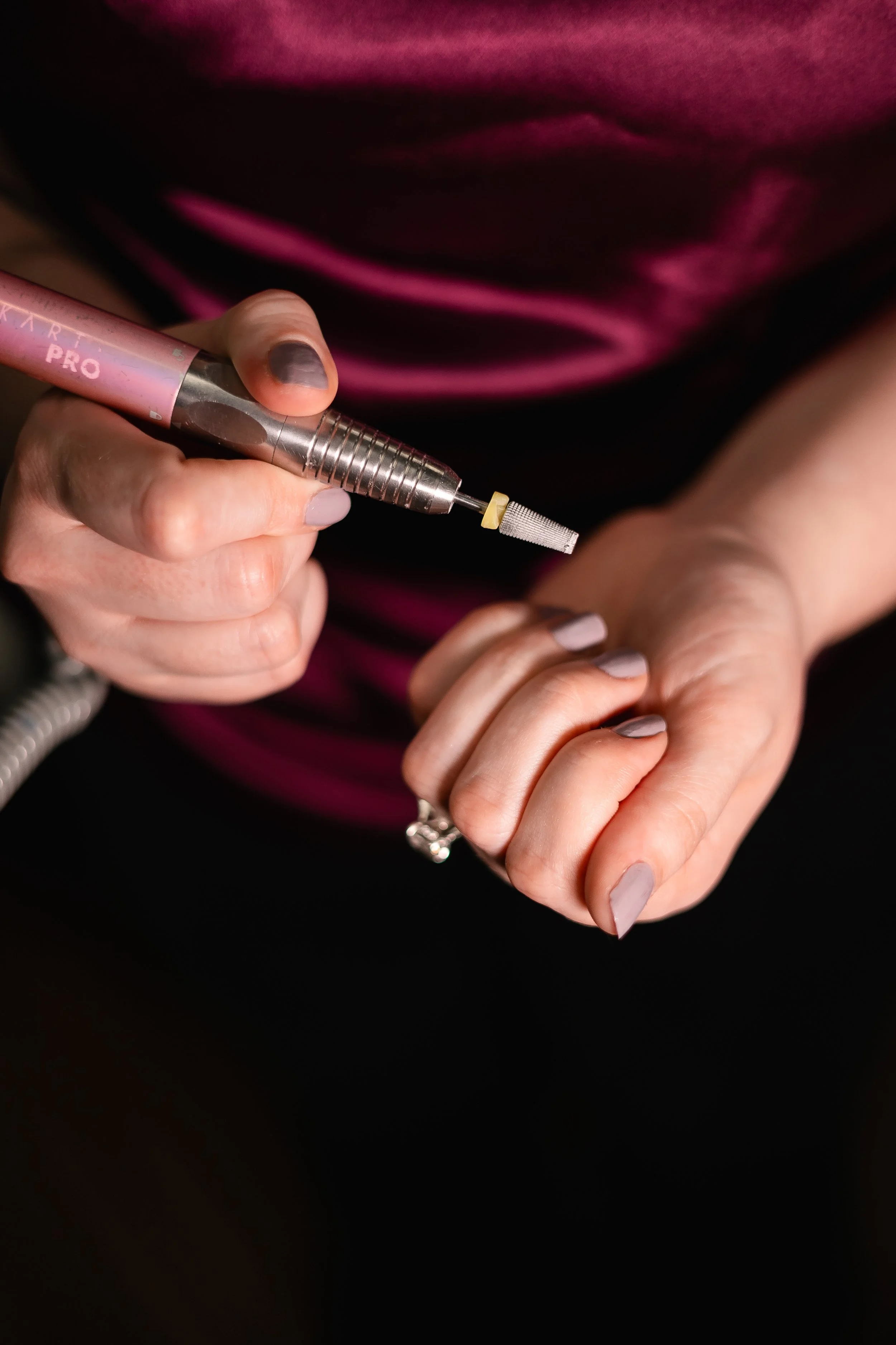 A person holding a small electronic device with a drill-like attachment in one hand, and their other hand with manicured nails. The person is wearing a ring and a dark purple shirt.