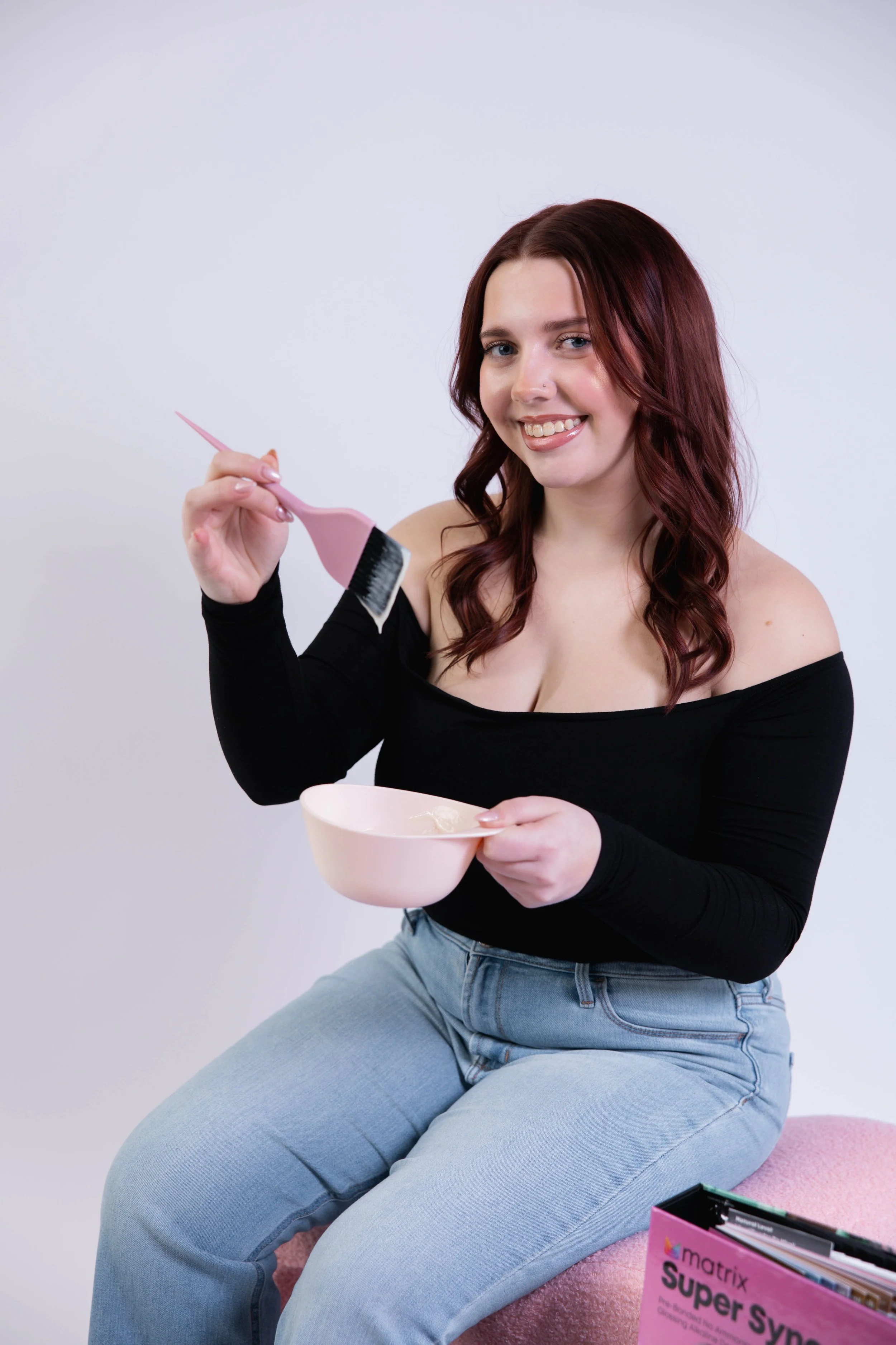 A young woman with red hair and a black off-the-shoulder top sitting on a pink stool, holding a pink bowl and a paintbrush, smiling at the camera, with a pink folder labeled 'Matrix Super Syn' nearby.