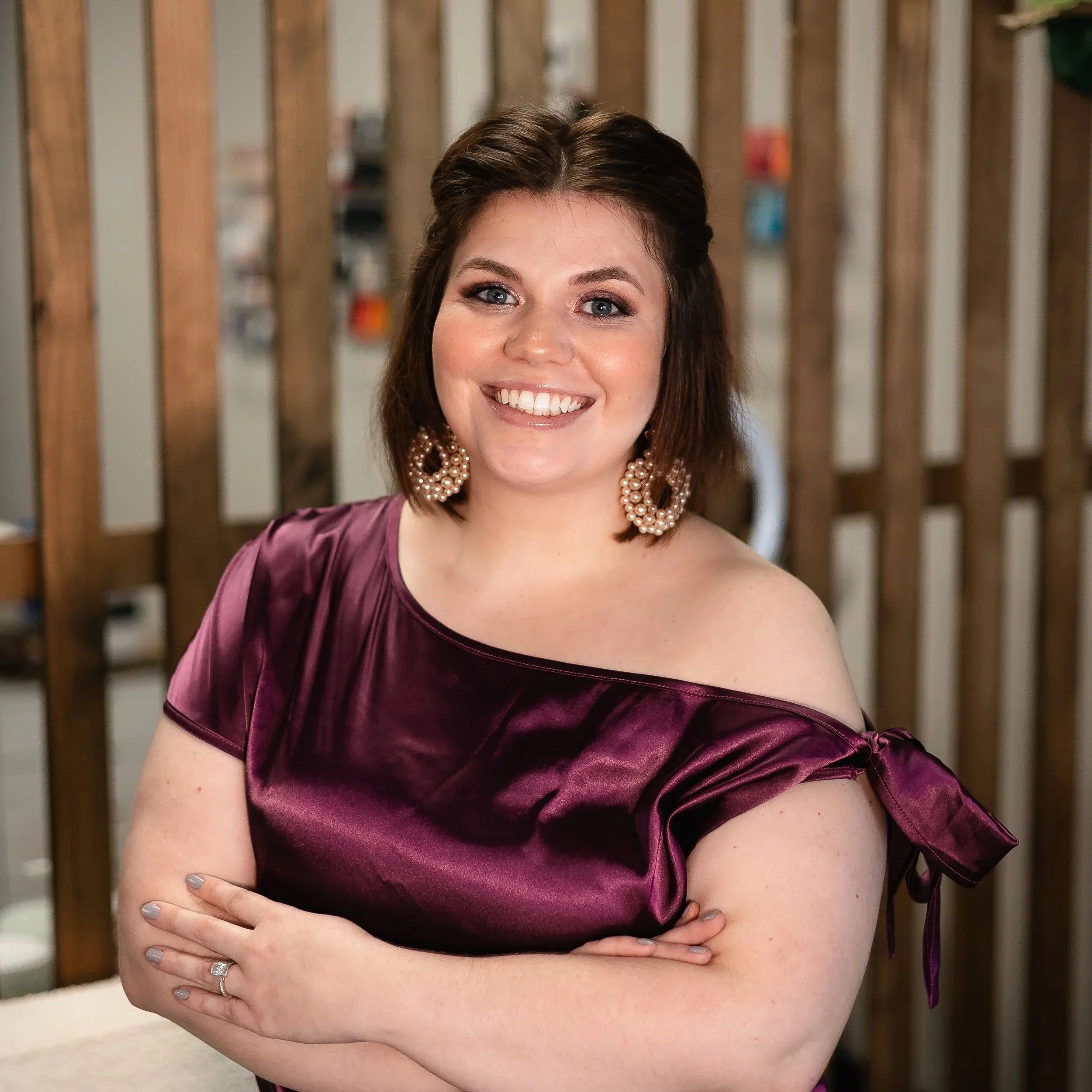 A woman with shoulder-length brown hair smiling, wearing a purple satin off-shoulder top with a bow on the shoulder and large pearl earrings, in front of a wooden divider.