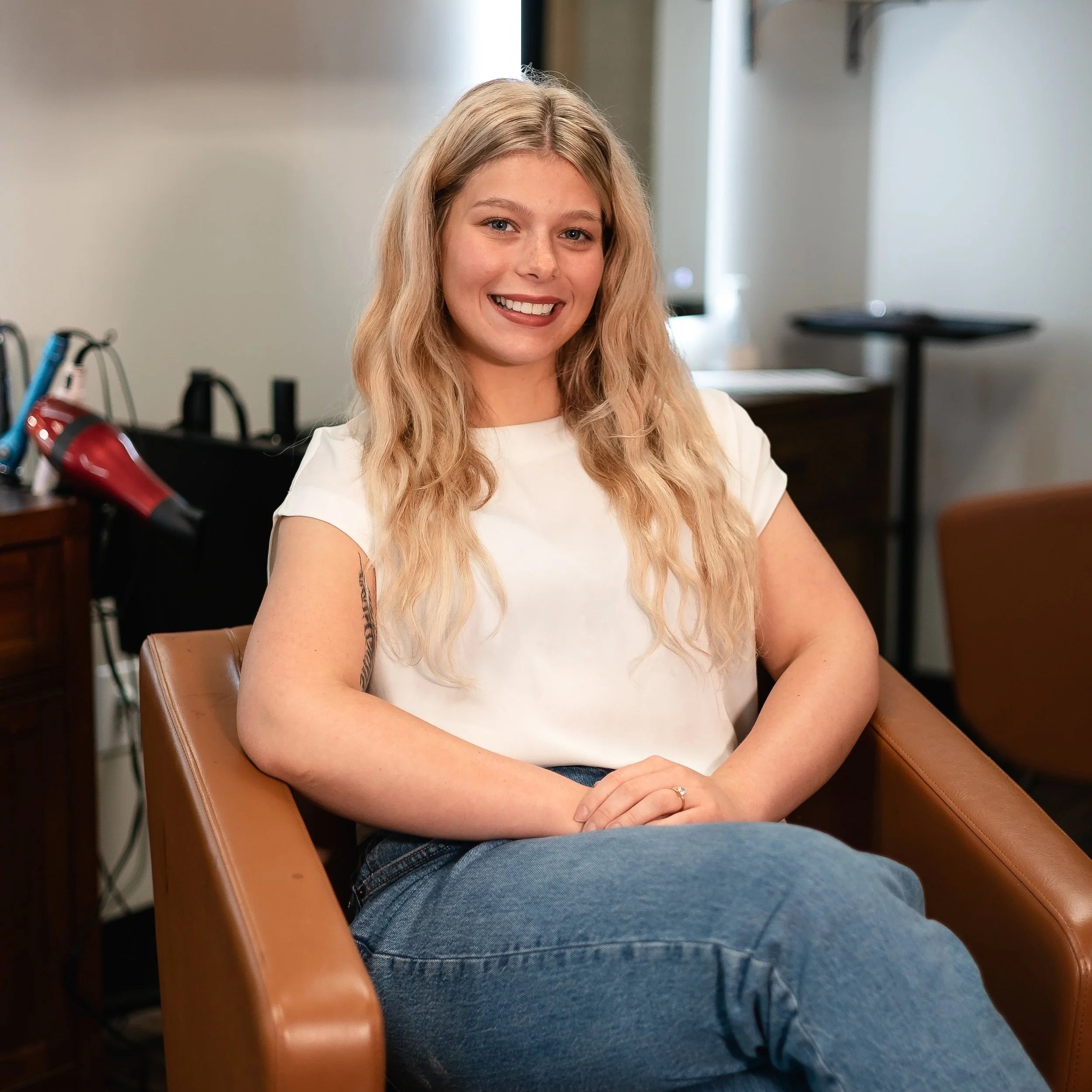 A young woman with long blonde wavy hair smiling and sitting in a brown leather chair in an indoor setting.