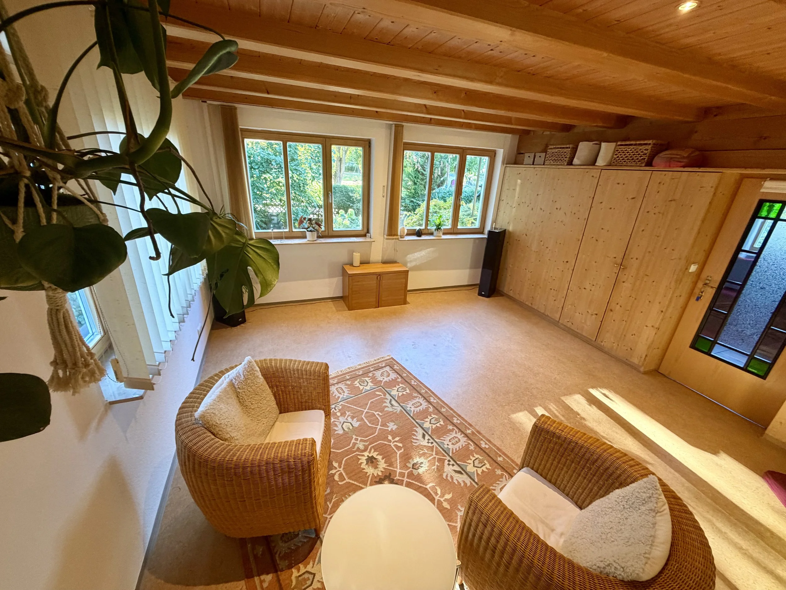 Ein gemütliches Wohnzimmer mit großen Fenstern, Holzwand- und Deckenverkleidung, einem kleinen Teppich, Rattanstühlen, einem kleinen Holzmöbel und Pflanzen