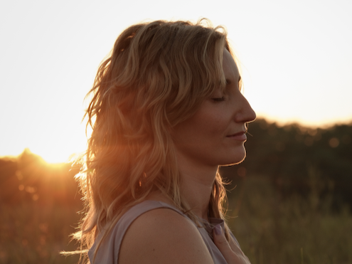 Eine Frau mit lockigem, blondem Haar steht im Sonnenuntergang in einer Naturumgebung.
