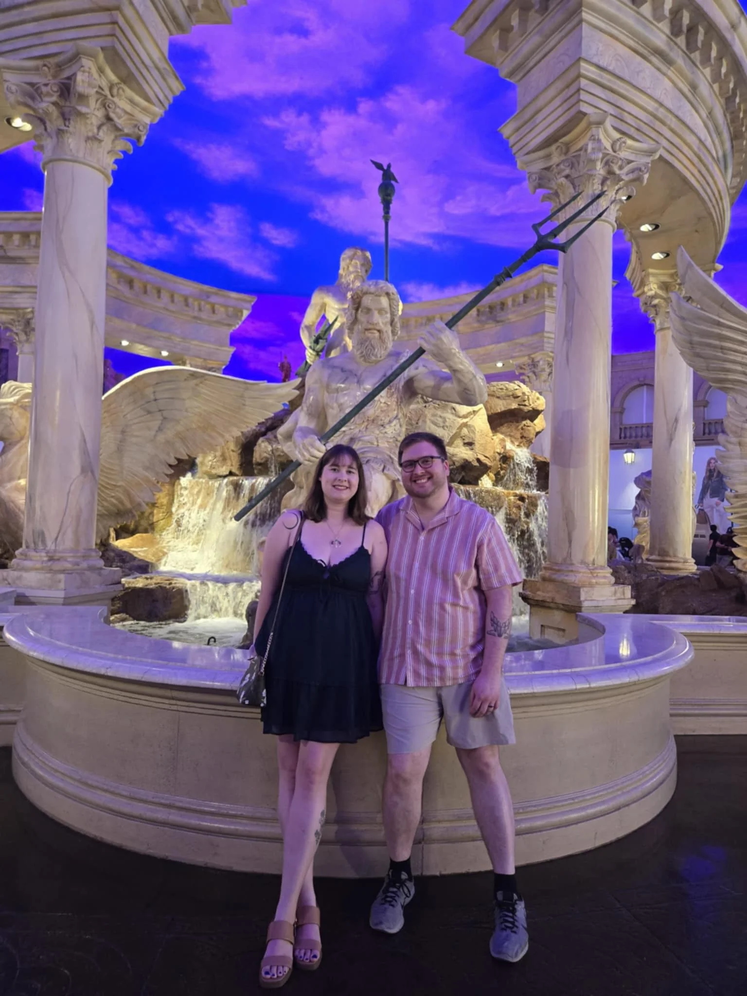 A young woman and a young man standing in front of a large, ornate fountain with statues, columns, and a blue sky background at a themed indoor location.
