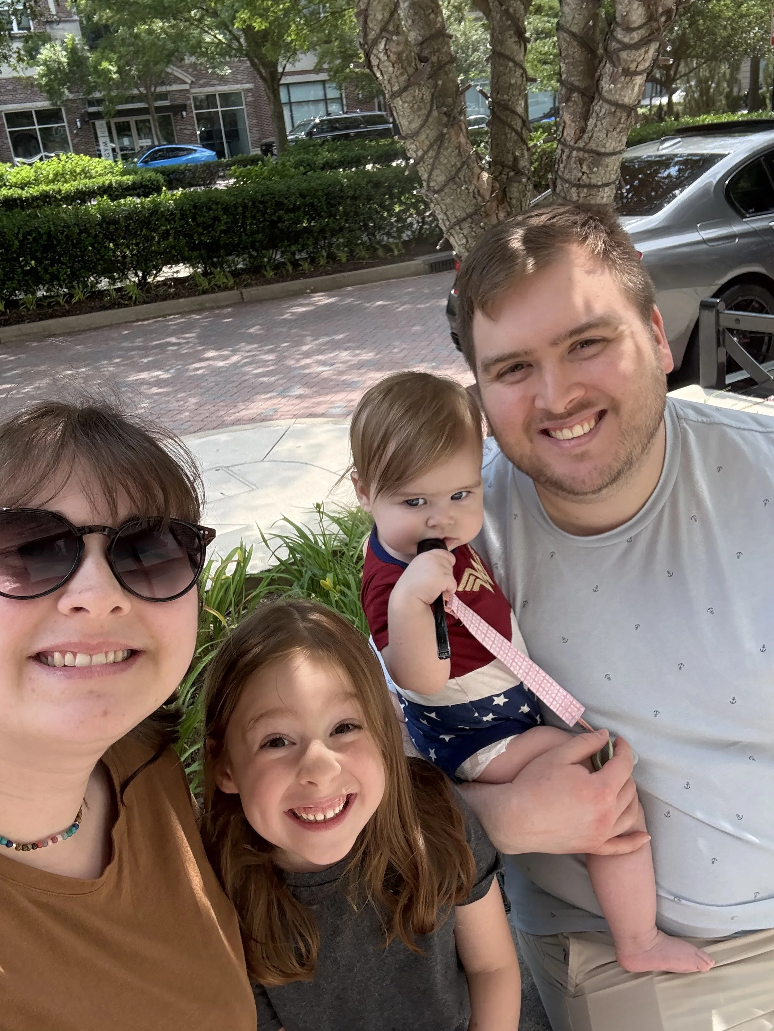 Family of four taking a selfie outdoors on a sunny day, with a woman wearing sunglasses, a young girl with red hair, a toddler dressed in Wonder Woman outfit, and a man smiling.