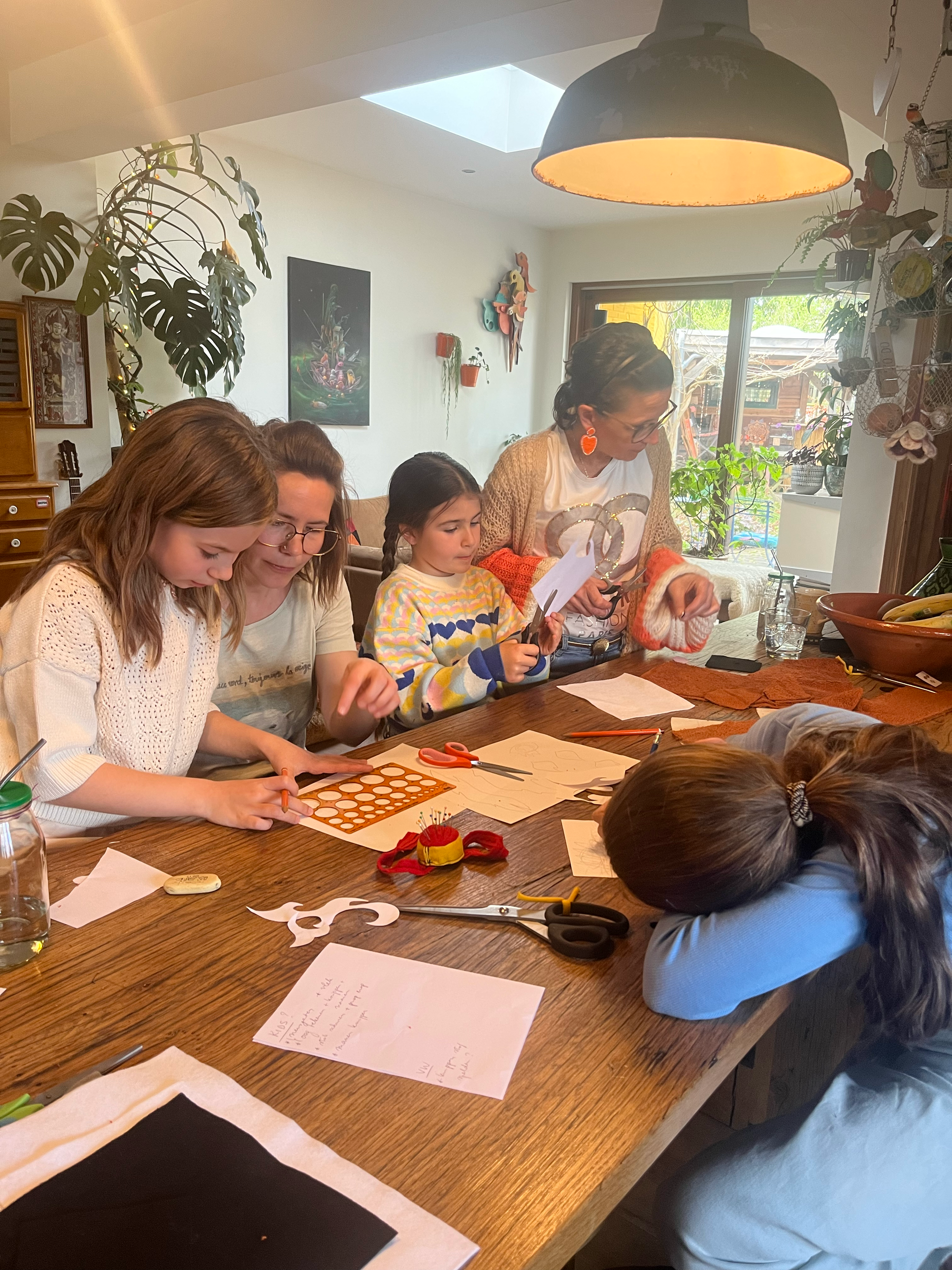 Een groep mensen rond een tafel doet een knutselactiviteit met papier en scharen in een huiselijke omgeving.