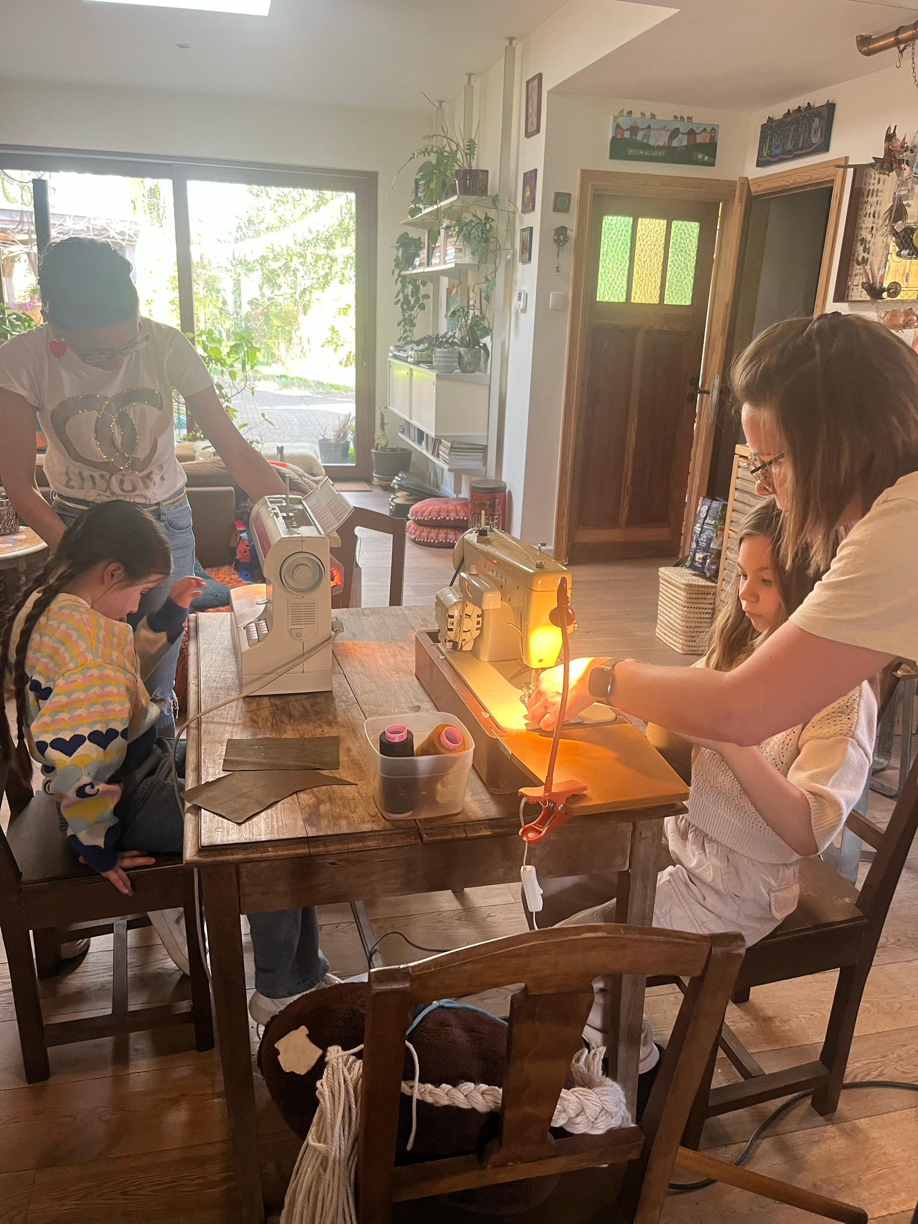 Twee personen werken samen aan een project op een houten tafel in een huiselijke omgeving. Er is een naaimachine, stoffen en knutselbenodigdheden zichtbaar. De ruimte heeft houten meubels en veel natuurlijk licht door grote ramen. Planten en decorati
