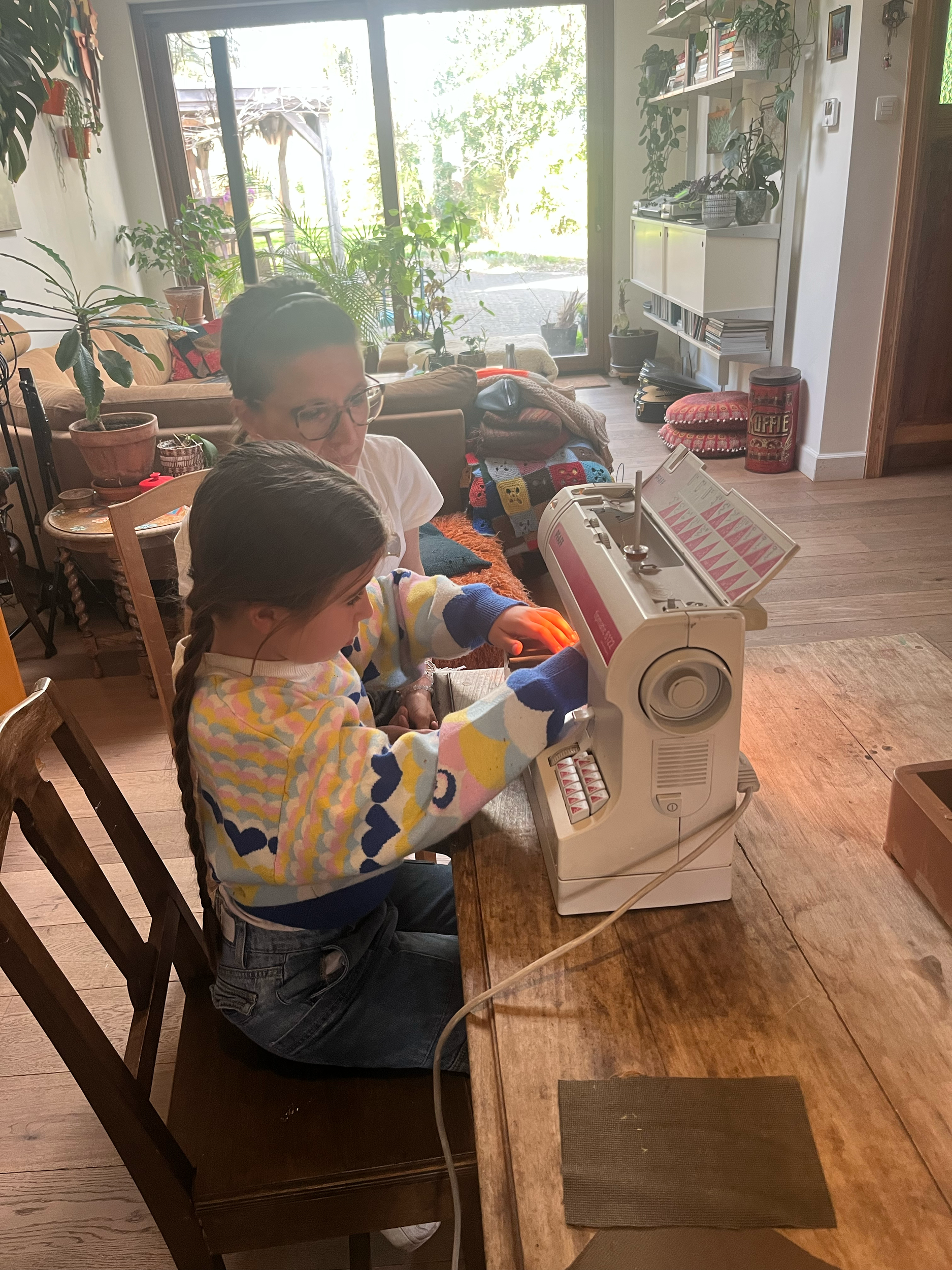 Twee personen gebruiken een naaimachine in een huiselijke omgeving, met veel planten en een grote raam.