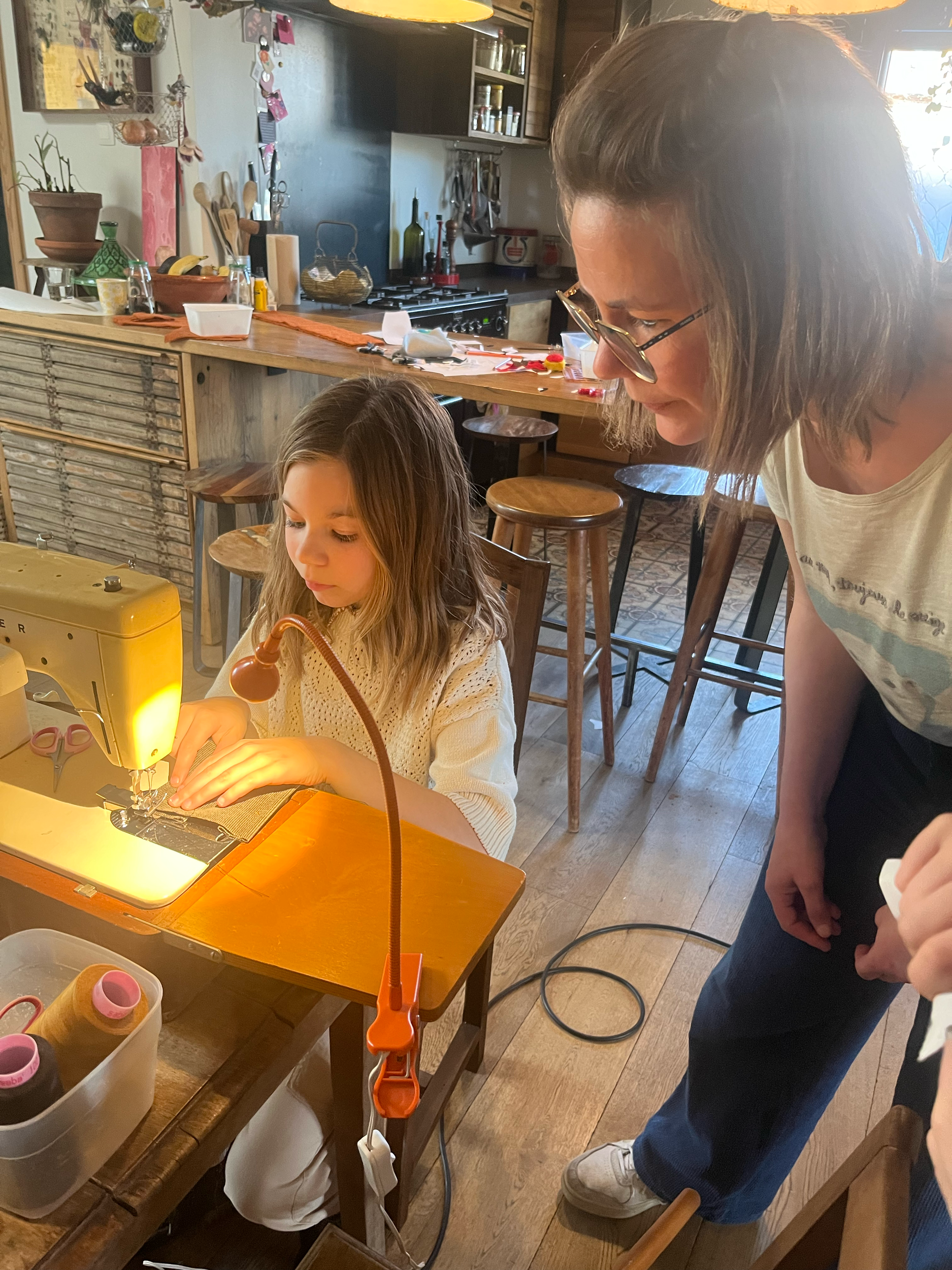 Twee mensen in een keuken, één met een naaimachine bezig, de ander kijkt toe. De omgeving is huiselijk met houten meubels en keukengerei op de achtergrond.