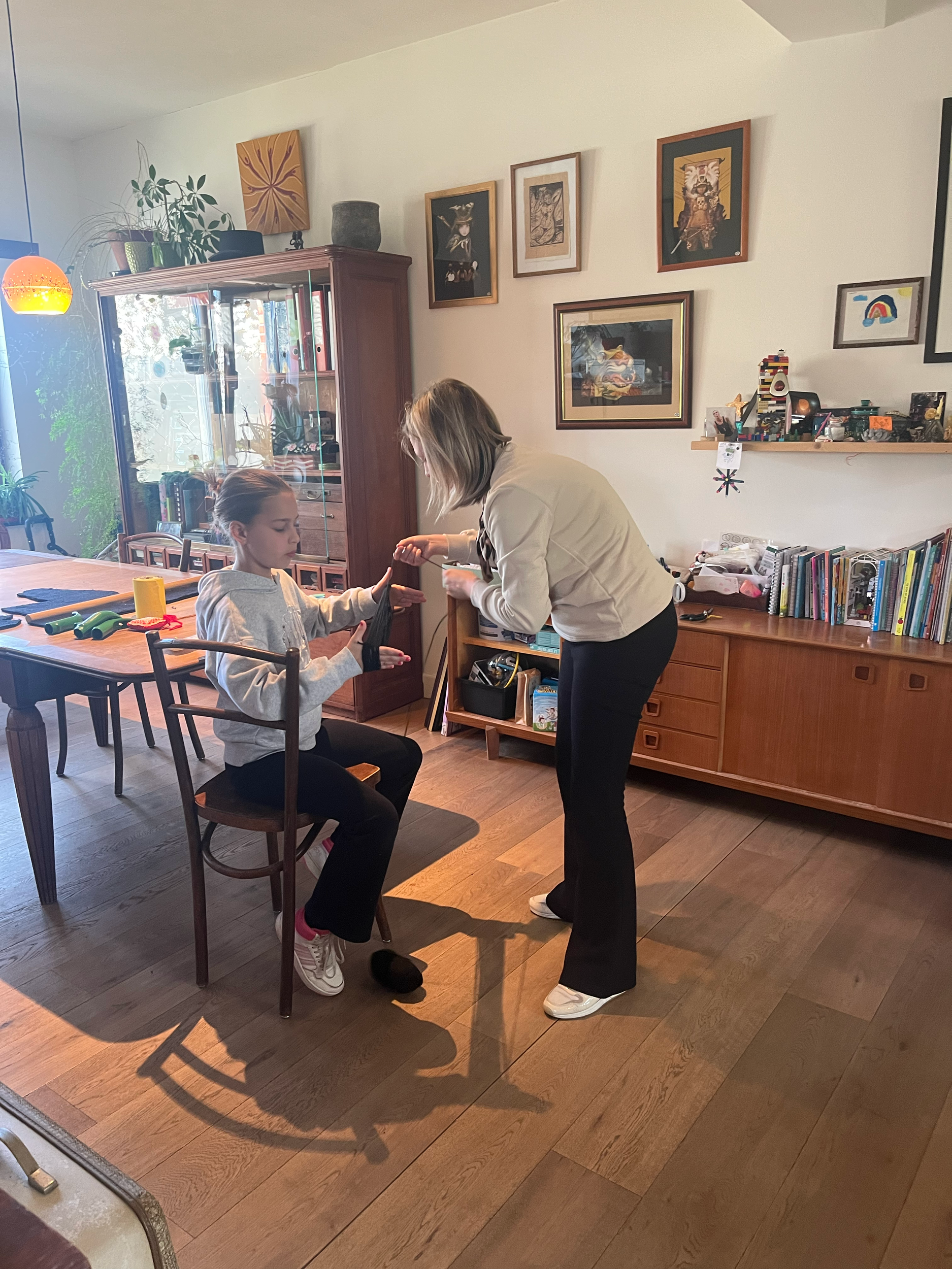 Twee personen in een woonkamer. Een persoon staat en lijkt iets aan de persoon die zit te geven. Er zijn boeken en kunst aan de muur.