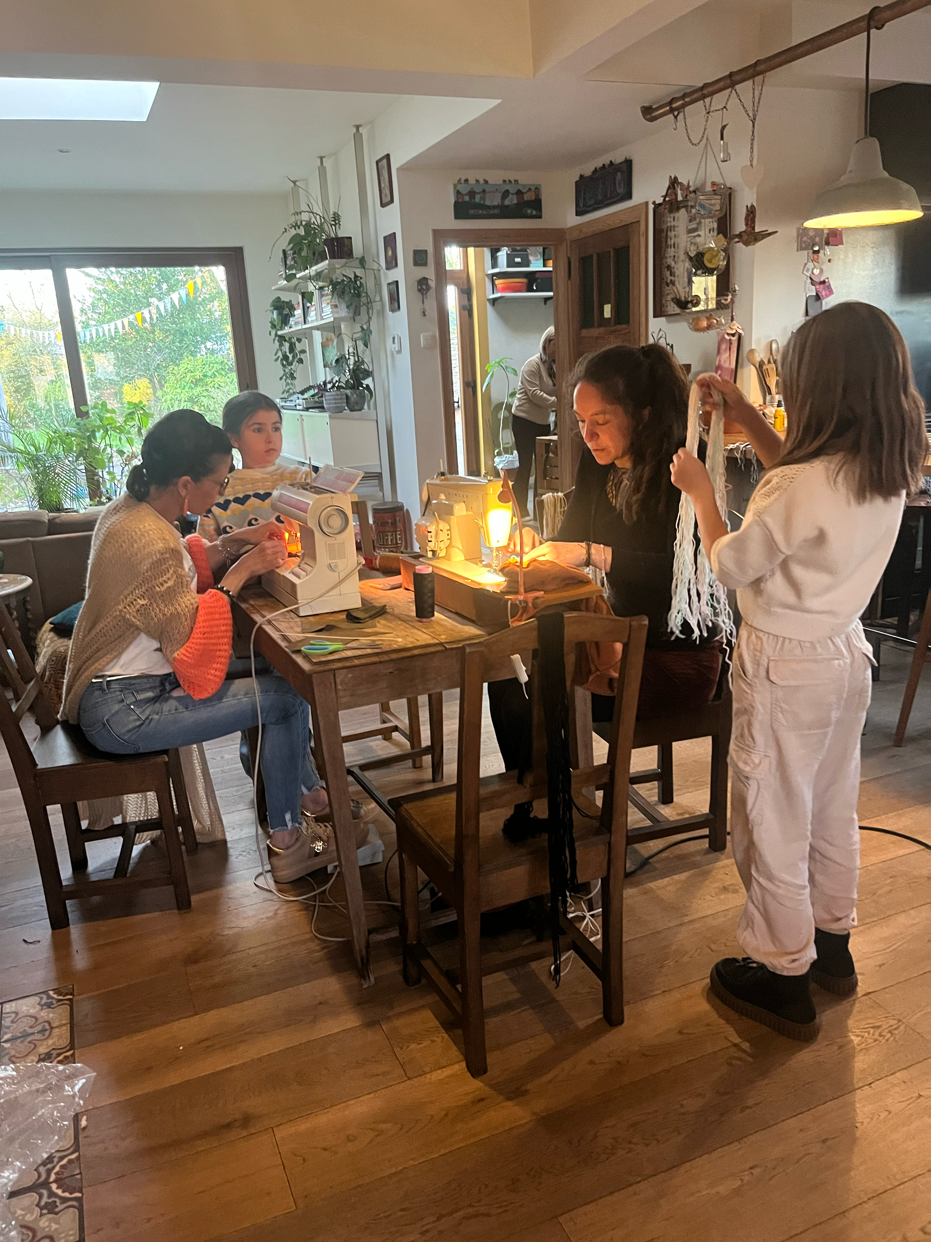 Groep mensen bezig met naaien, zittend aan een tafel in een huiselijke omgeving. Er zijn naaimachines en stoffen op de tafel, en een kind staat en kijkt toe. De kamer is ingericht met planten en decoratieve items.