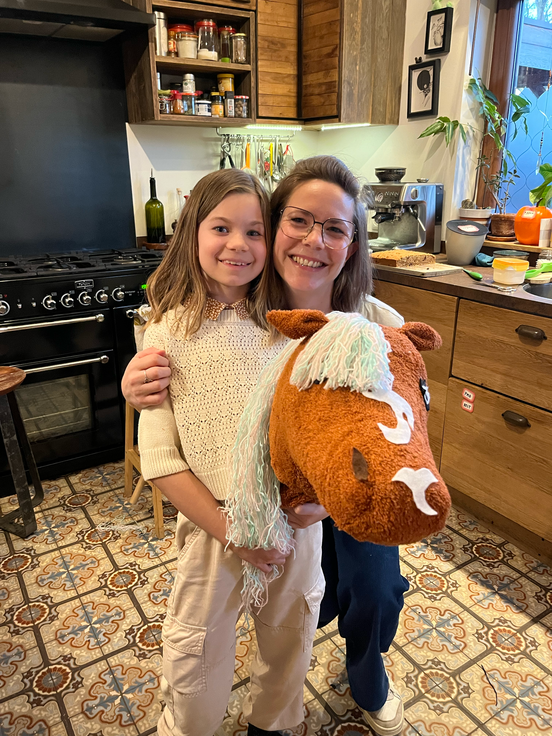 Twee personen poseren samen in een keuken, waarbij een van hen een grote speelgoedknuffel vasthoudt. De keuken heeft een retro tegelvloer en houten kasten met verschillende potjes ingrediënten.