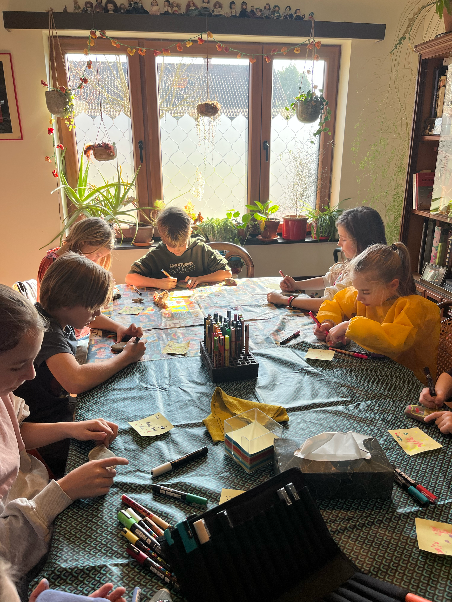 Groep jonge mensen die aan een tafel zit te knutselen, met pennen, stiften en papier. De tafel is bedekt met een blauw tafelkleed en er zijn planten bij het raam.
