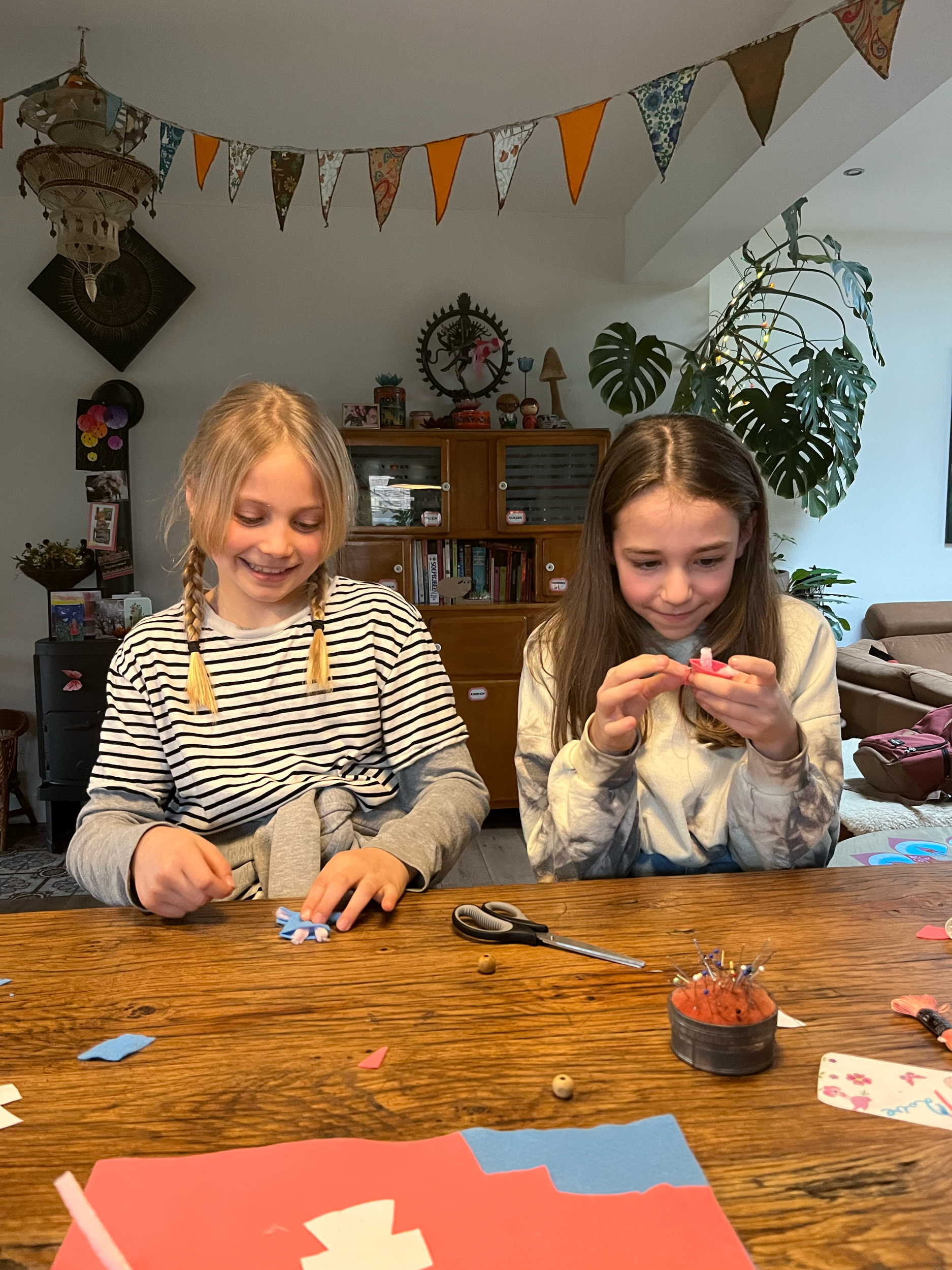 Twee kinderen knutselen aan een tafel met gekleurd papier, schaar en plakspullen. De ruimte is versierd met slingers en een grote plant op de achtergrond.