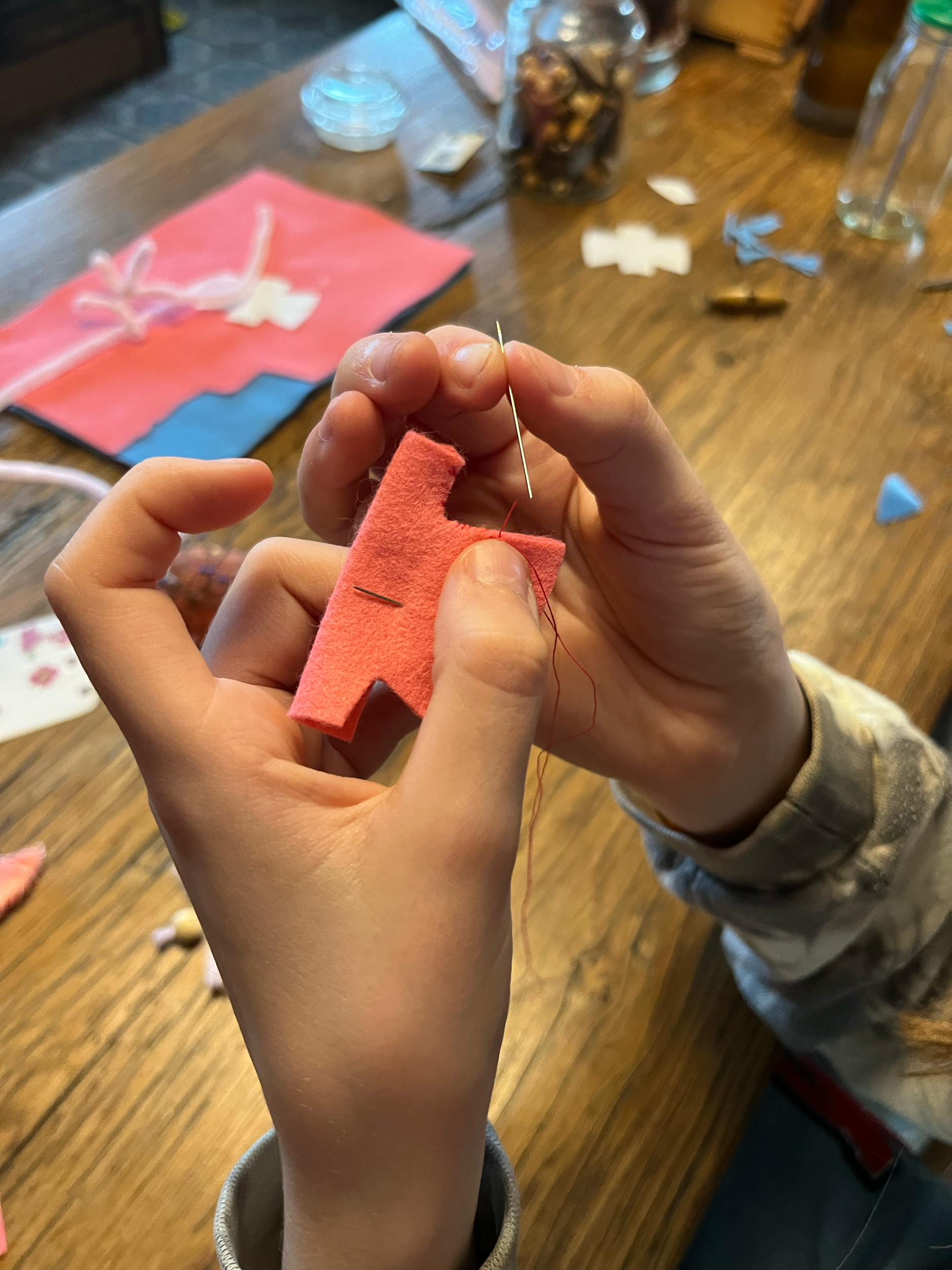Een paar handen die een rood stuk stof naaien met een naald en draad op een houten tafel met knutselspullen.