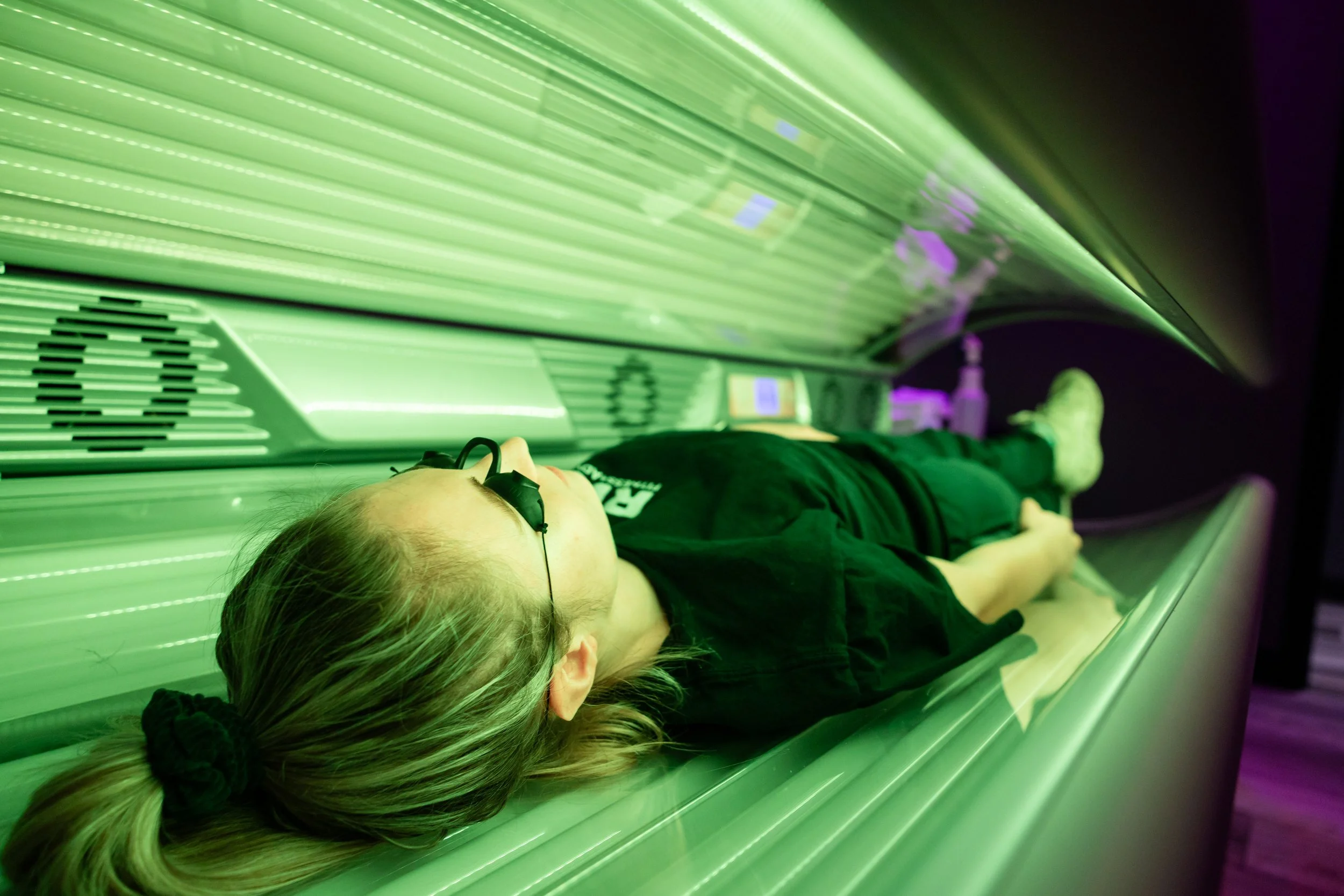 Person lying on a tanning bed under green UV light, wearing black clothing, glasses, with their hair in a ponytail.