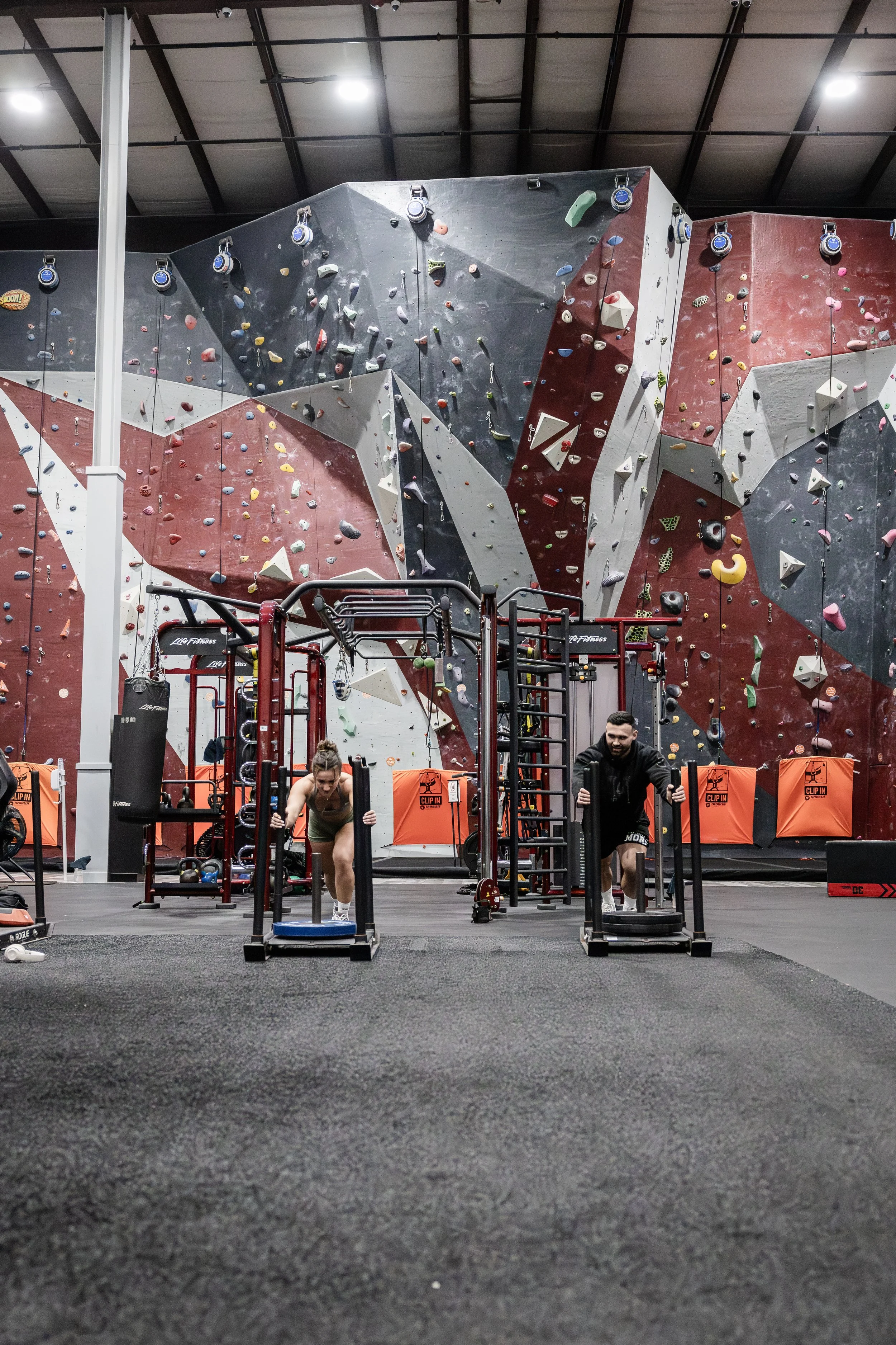 A gym with a climbing wall in the background and two people working out with sled pushes in the foreground.