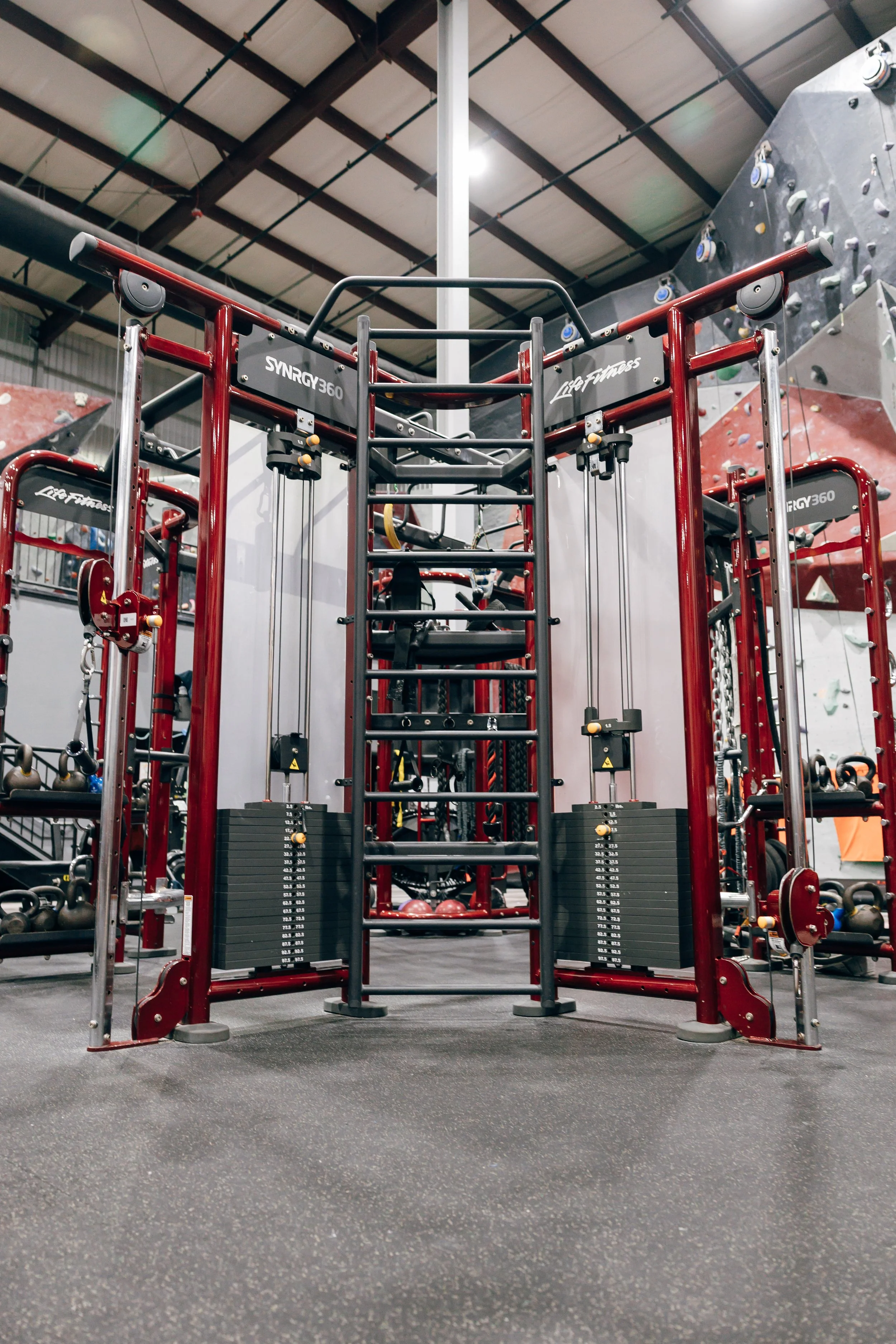 Inside a gym with a red and black multi-functional workout machine, weight plates, kettlebells, and a rock climbing wall in the background.