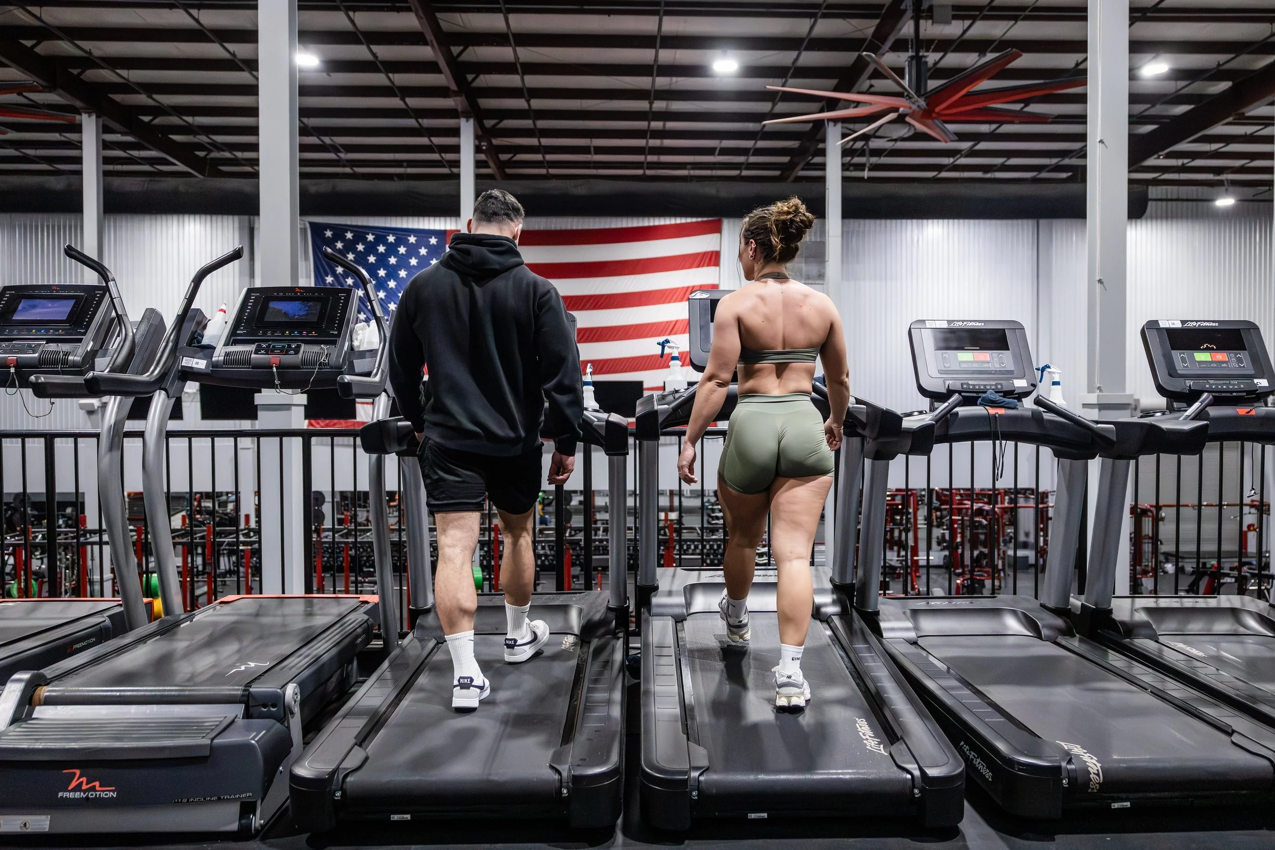 Two people walking on treadmills in a gym with an American flag in the background.