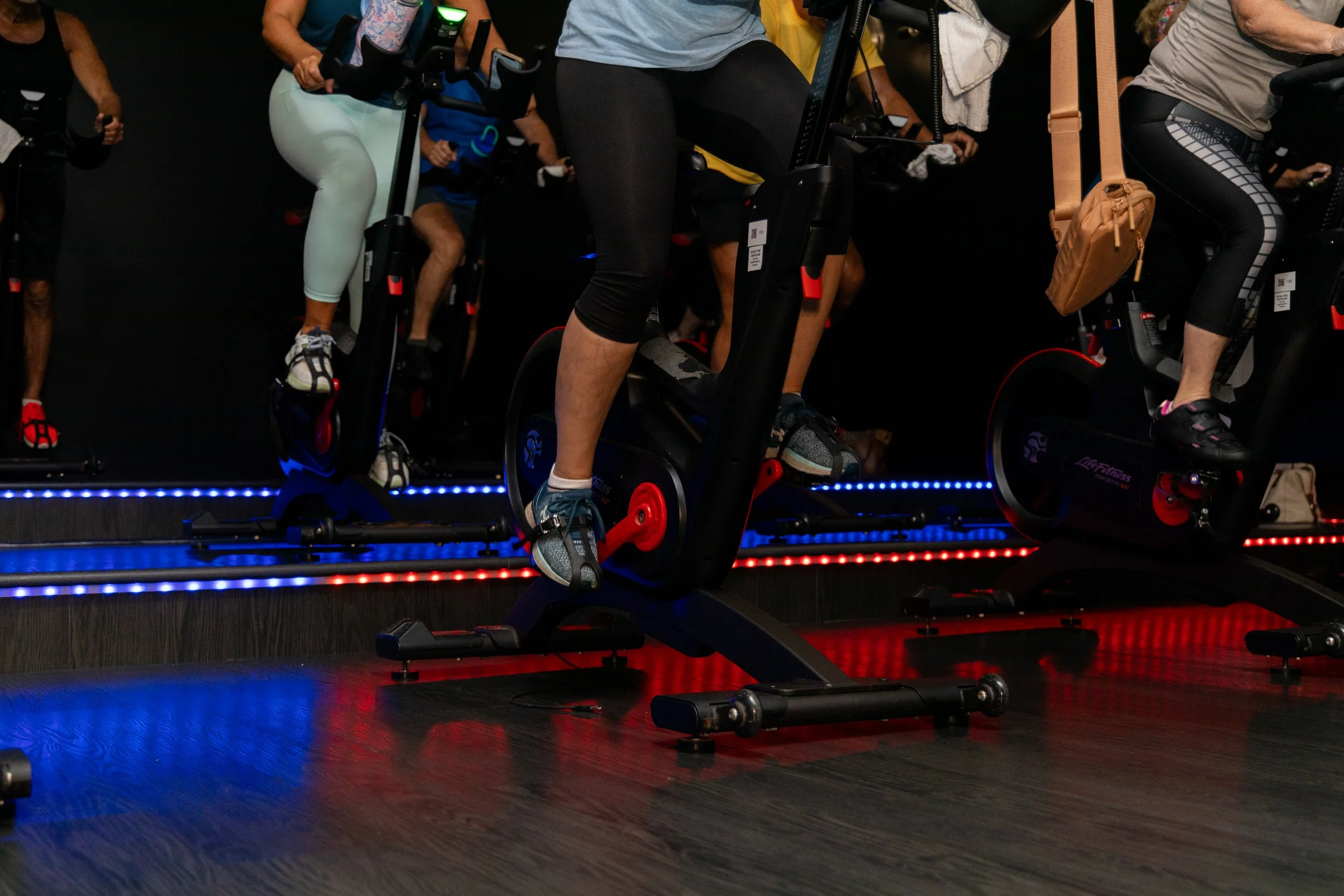 A woman practicing kickboxing at a gym, wearing blue boxing gloves and kicking a padded focus mitt.