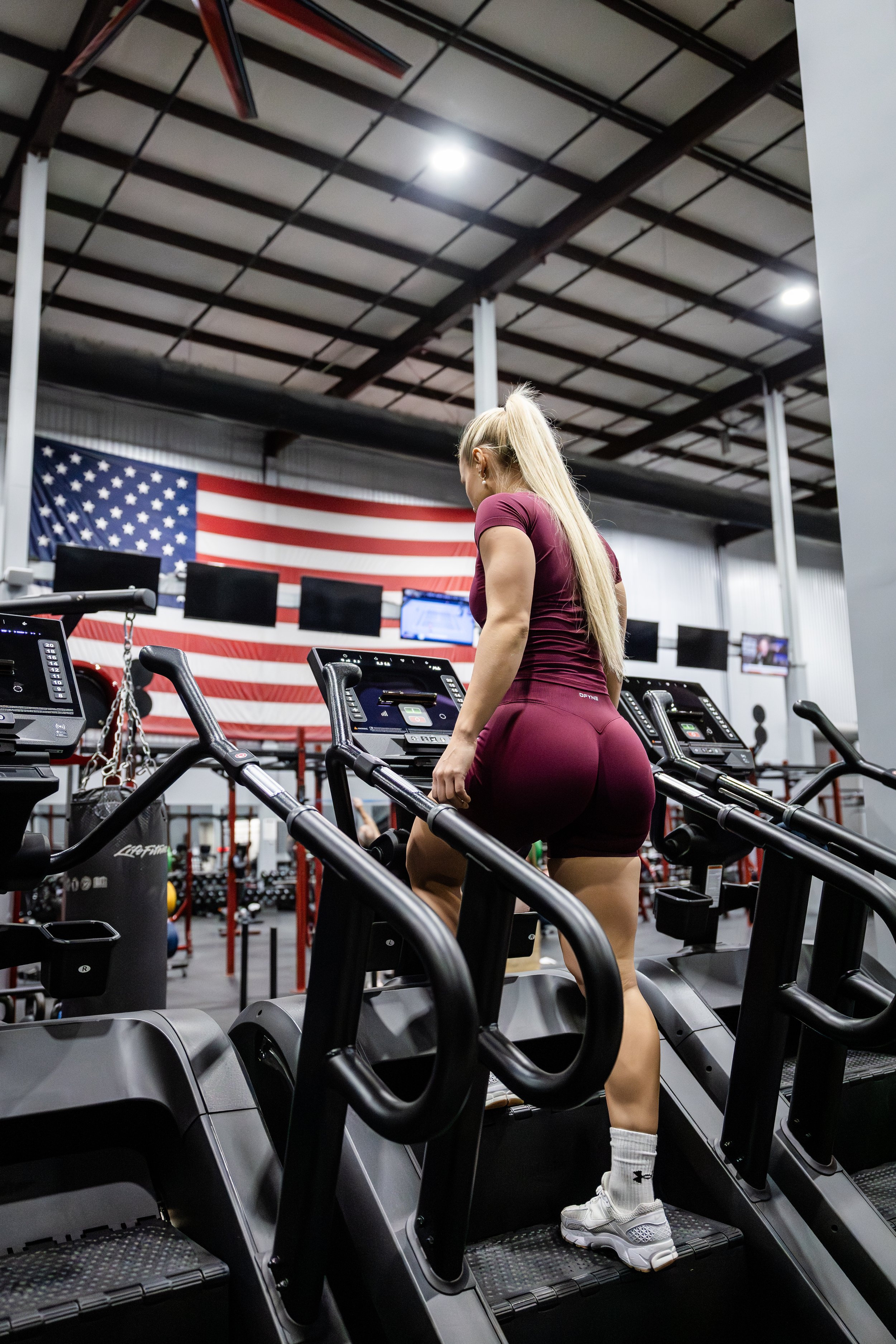 A woman with long blonde hair working out on a stair climbing machine inside a gym with an American flag on the wall.