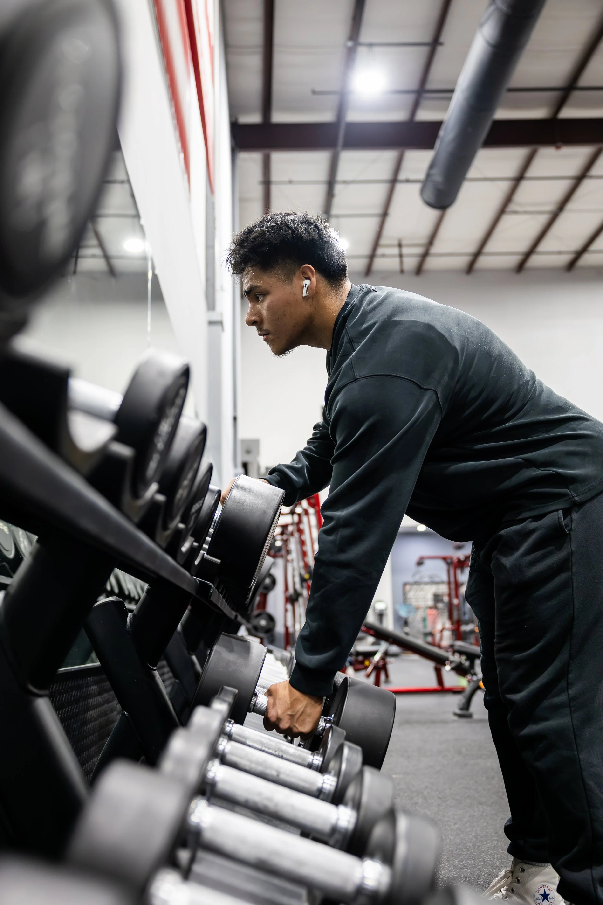 A man in black workout clothes lifting a dumbbell in a gym with racks of dumbbells and exercise equipment in the background.