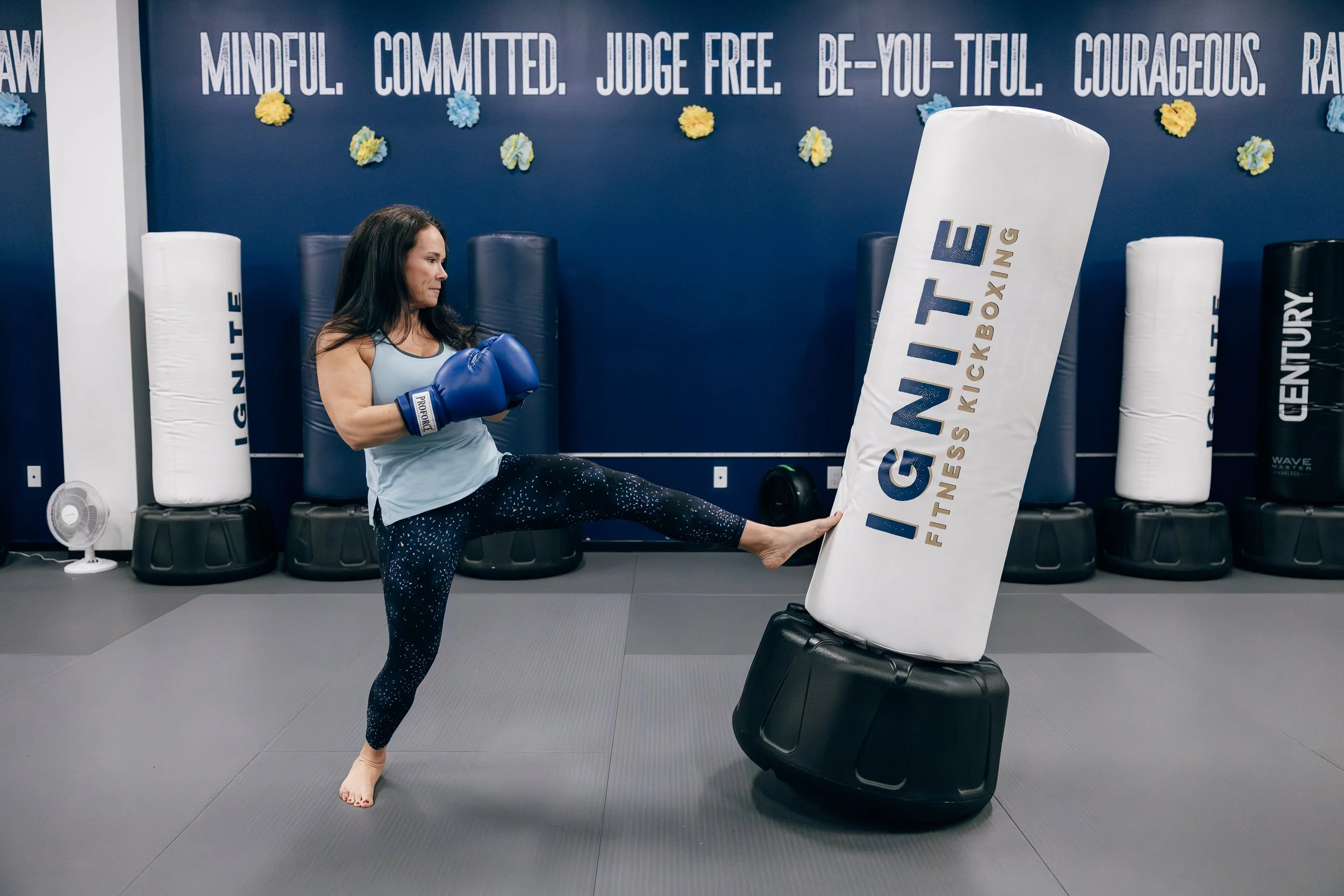 A woman practicing kickboxing at a gym, wearing blue boxing gloves and kicking a padded focus mitt.