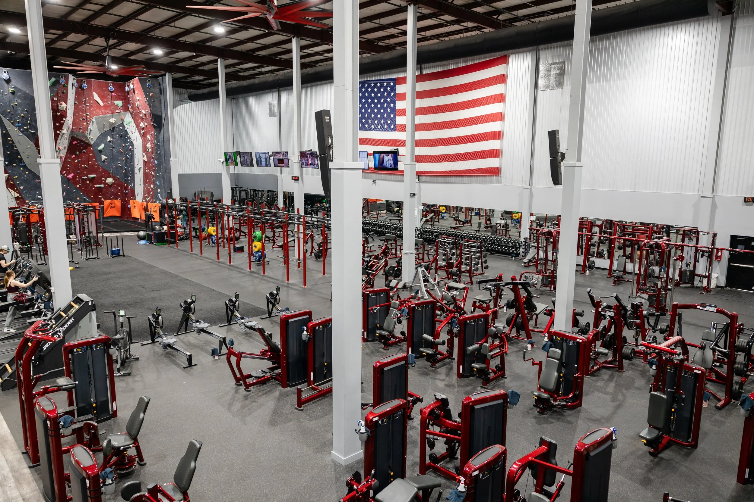 Inside a gym with a red and black multi-functional workout machine, weight plates, kettlebells, and a rock climbing wall in the background.