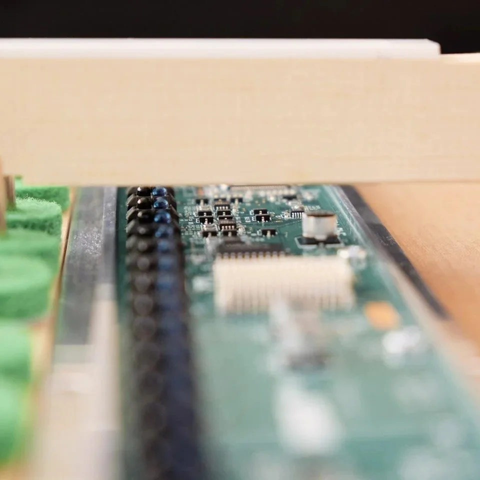 Nahaufnahme einer grünen Leiterplatte mit elektronischen Bauteilen, teils verdeckt durch eine Holzplatte.