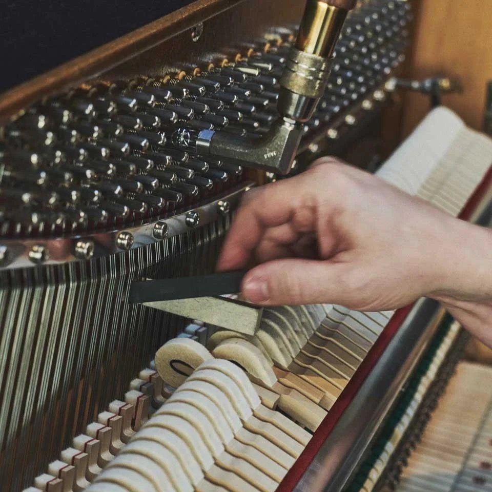 Nahaufnahme eines Fingers, der einen Keil zum Einstellen eines Klavierspiels in die Mechanik eines Klaviers steckt, mit sichtbaren Saiten und Hämmer im Inneren.