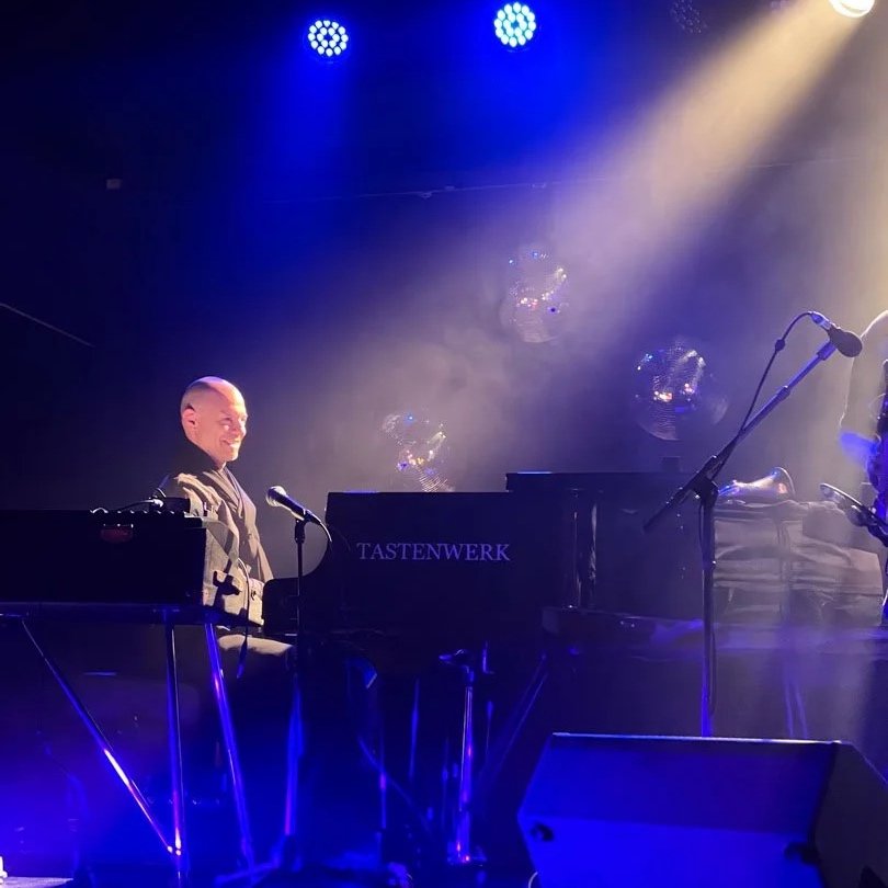 Ein Musiker spielt Klavier auf einer Bühne mit blauer Beleuchtung, im Hintergrund sind reflektierende Discokugeln und Nebel.
