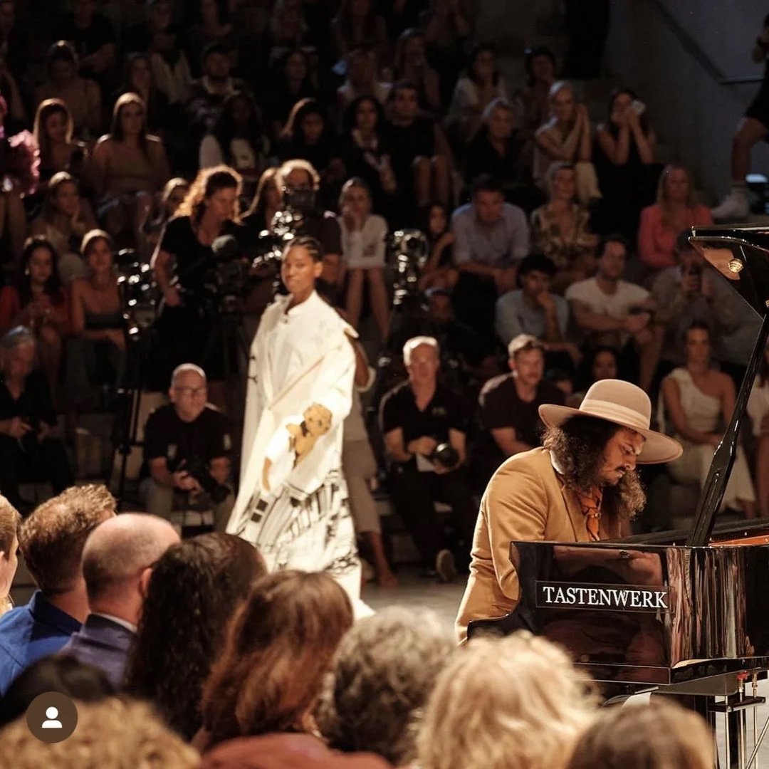 Musiker spielt Klavier während einer Bühnenshow vor einem Publikum, im Hintergrund eine Frau in einem weißen Kleid mit Teddy in der Hand. Viele Zuschauer sitzen und schauen in Richtung Bühne, einige filmen die Performance.