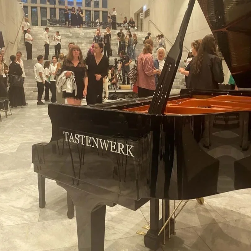 Schwarz glänzender Flügel mit der Aufschrift "TASTENWERK" in einer beliebten Einrichtung, umgeben von Menschen, die sich unterhalten, manche sitzen auf den Treppen im Hintergrund.