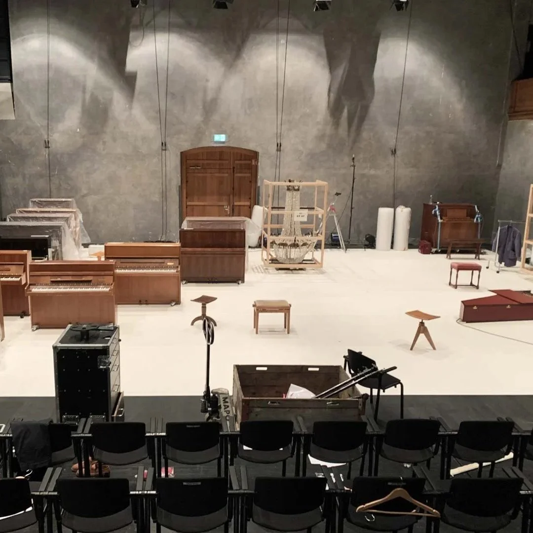 Ein leerer Theater- oder Konzertsaal mit verschiedenen Musikinstrumenten und Bühnenrequisiten, darunter Klaviere, Stühle, Lampen und ein Kristalllob.