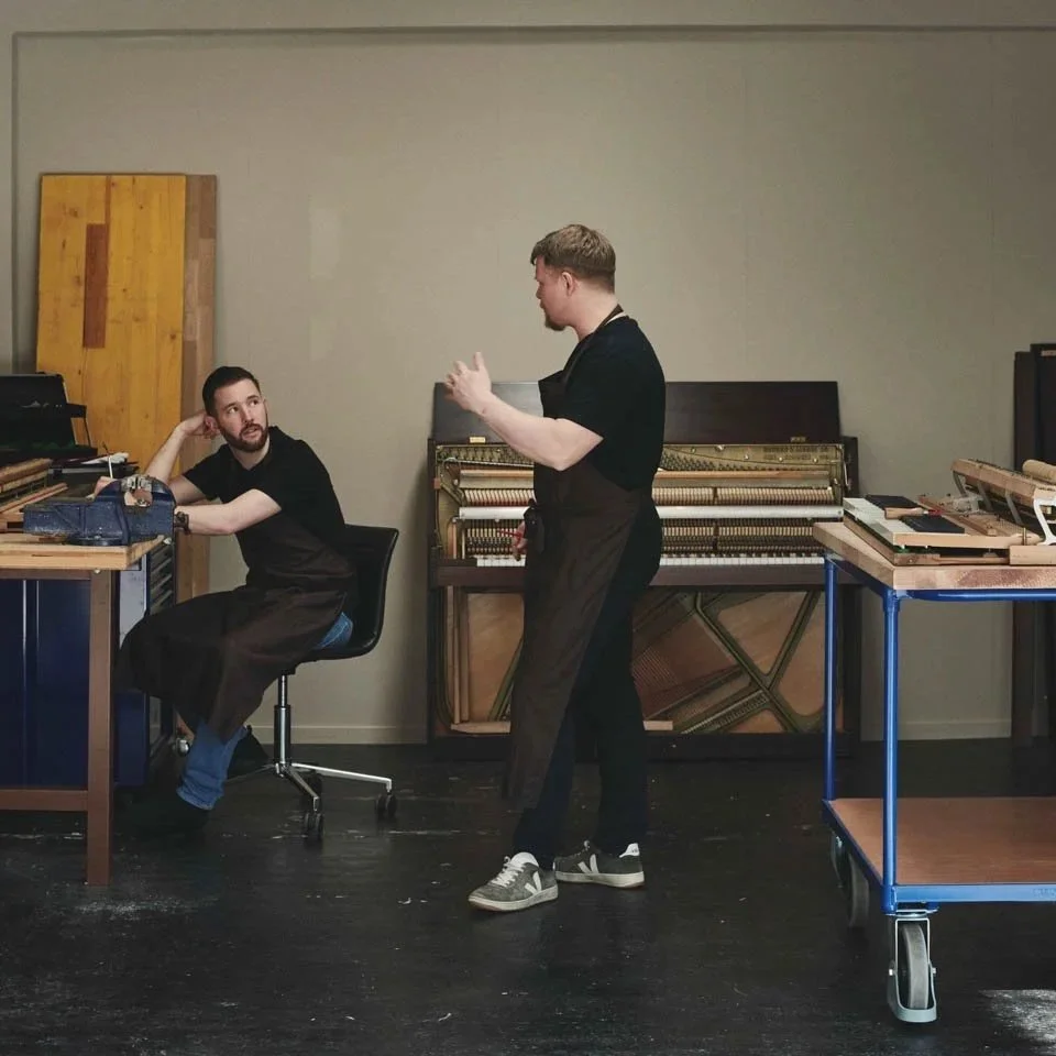 Stefan Blumer und Silvan Stürzinger in einem Musikraum, einer sitzt an einem Tisch mit einem Klavier, der andere steht und diskutiert. Im Hintergrund befinden sich teilweise zerlegte Klaviere.