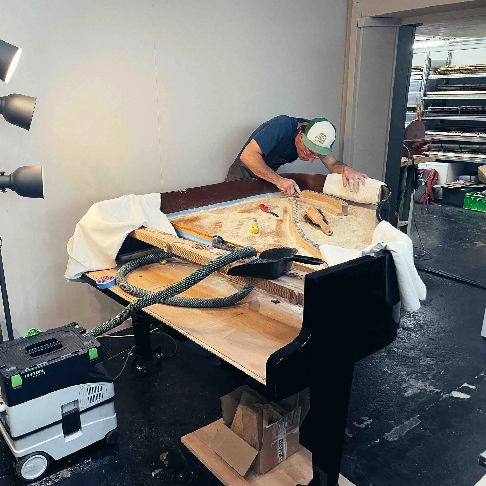 Flavio Morganti arbeitet an einem Flügel in einem Raum mit Werkstatteinrichtung. Es sind Werkzeuge und Zubehör auf einem Tisch, und im Hintergrund sind Regale mit Bau- und Werkzeugmaterial.