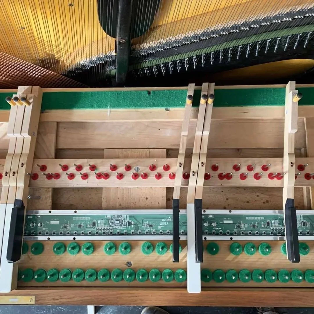 Innenaufnahme eines Klaviers mit sichtbarer Mechanik, darunter eine Holzplatte mit roten, grünen und schwarzen Komponenten, die Teile der Tasten- und Hämmermechanik darstellen.