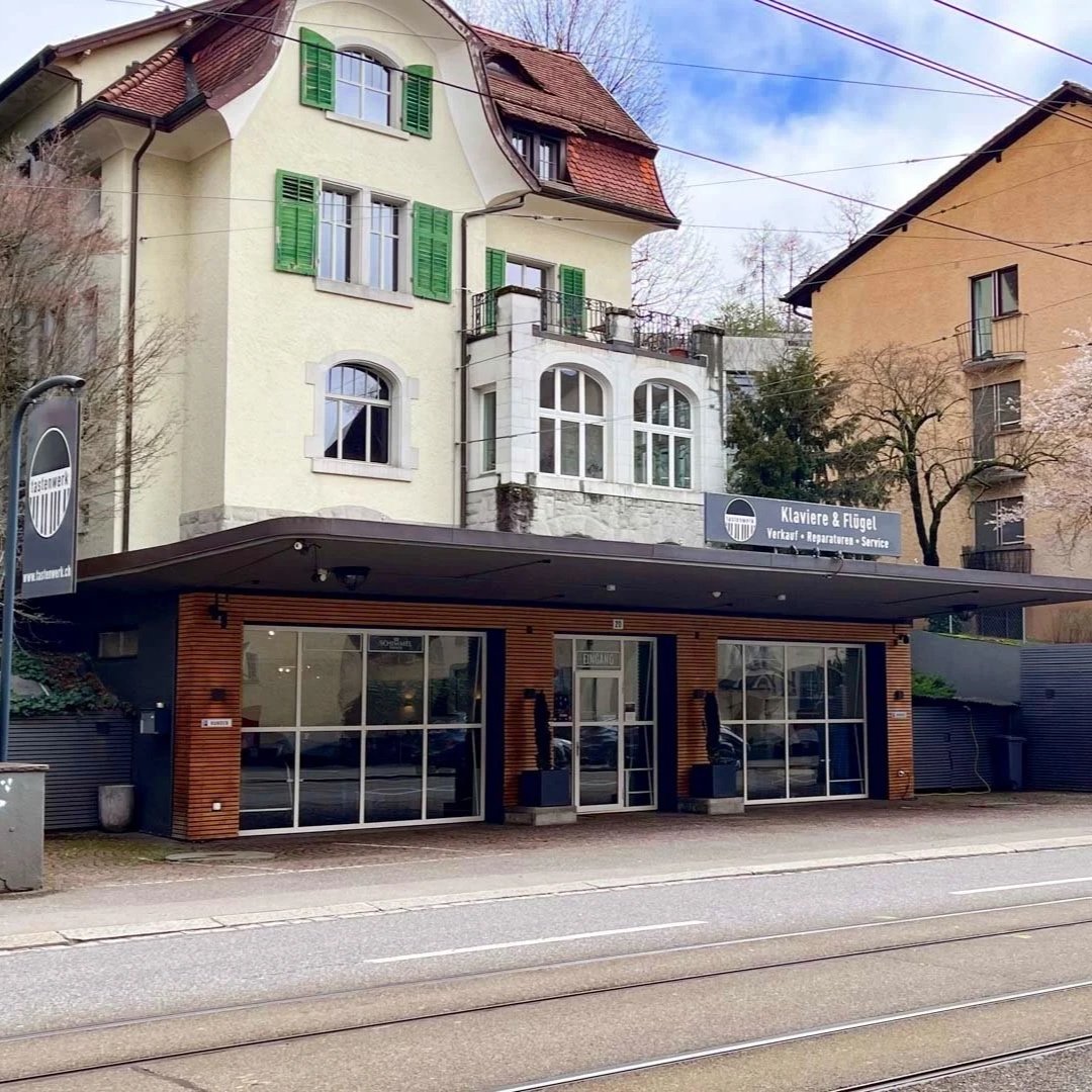 Ein modernes Geschäftsgebäude mit Glasfront und Holzverkleidung, das den Laden "Klavfere & Flügel" für Verkauf, Reparatur und Service von Klavieren und Flügeln beherbergt, in einer Straße mit Tramgleisen.