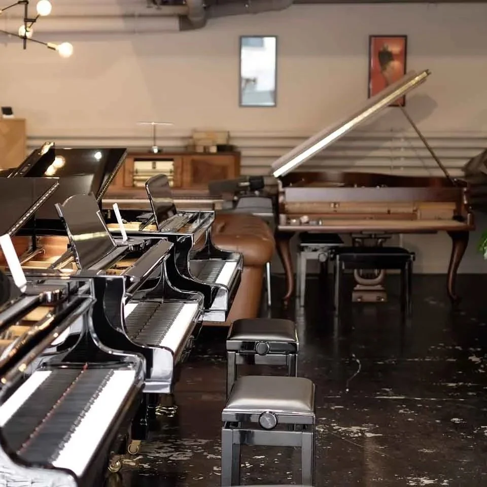 Mehrere Klaviere in einem Musikzimmer, eines mit offenem Deckel, auf Holzboden.