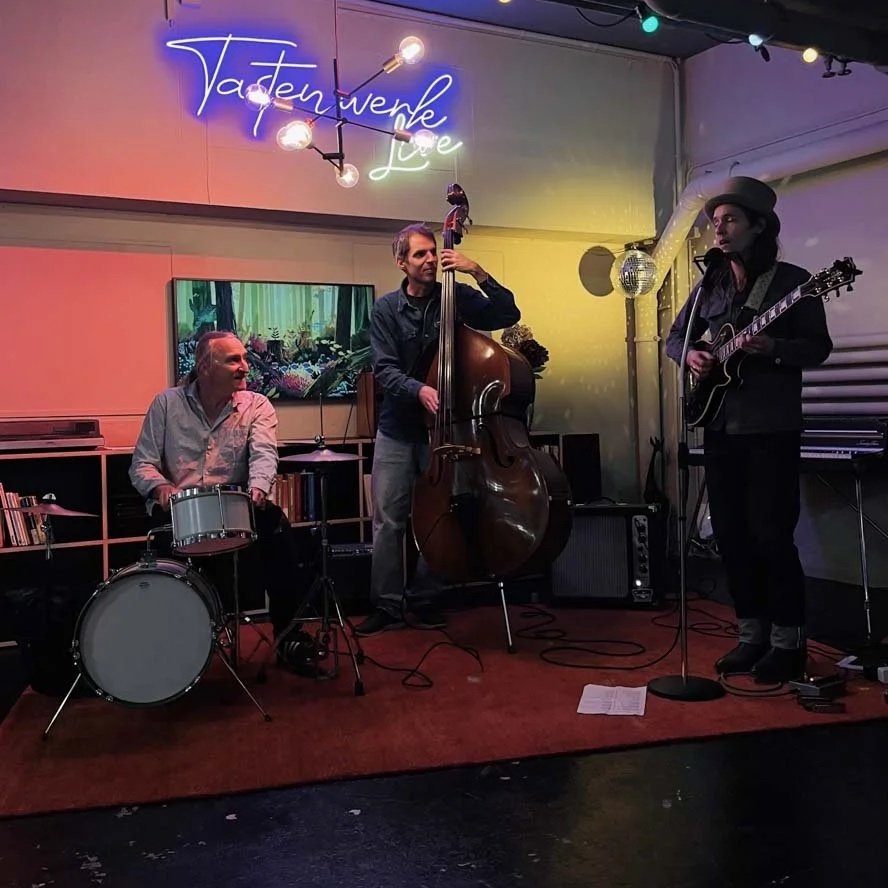 Drei Musiker spielen in einer Bar: ein Schlagzeuger, ein Kontrabassspieler und ein Gitarrist. Im Hintergrund sind bunte Lichter und eine Neon-Schrift, die 'Vaderweise Love' lautet.