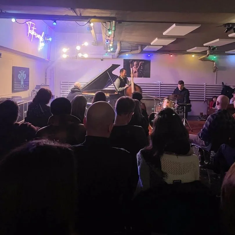 Jazzband mit Klavier, Kontrabass und Schlagzeug spielt vor Publikum in einem kleinen, dunklen Raum, der mit bunten Lichter dekoriert ist.