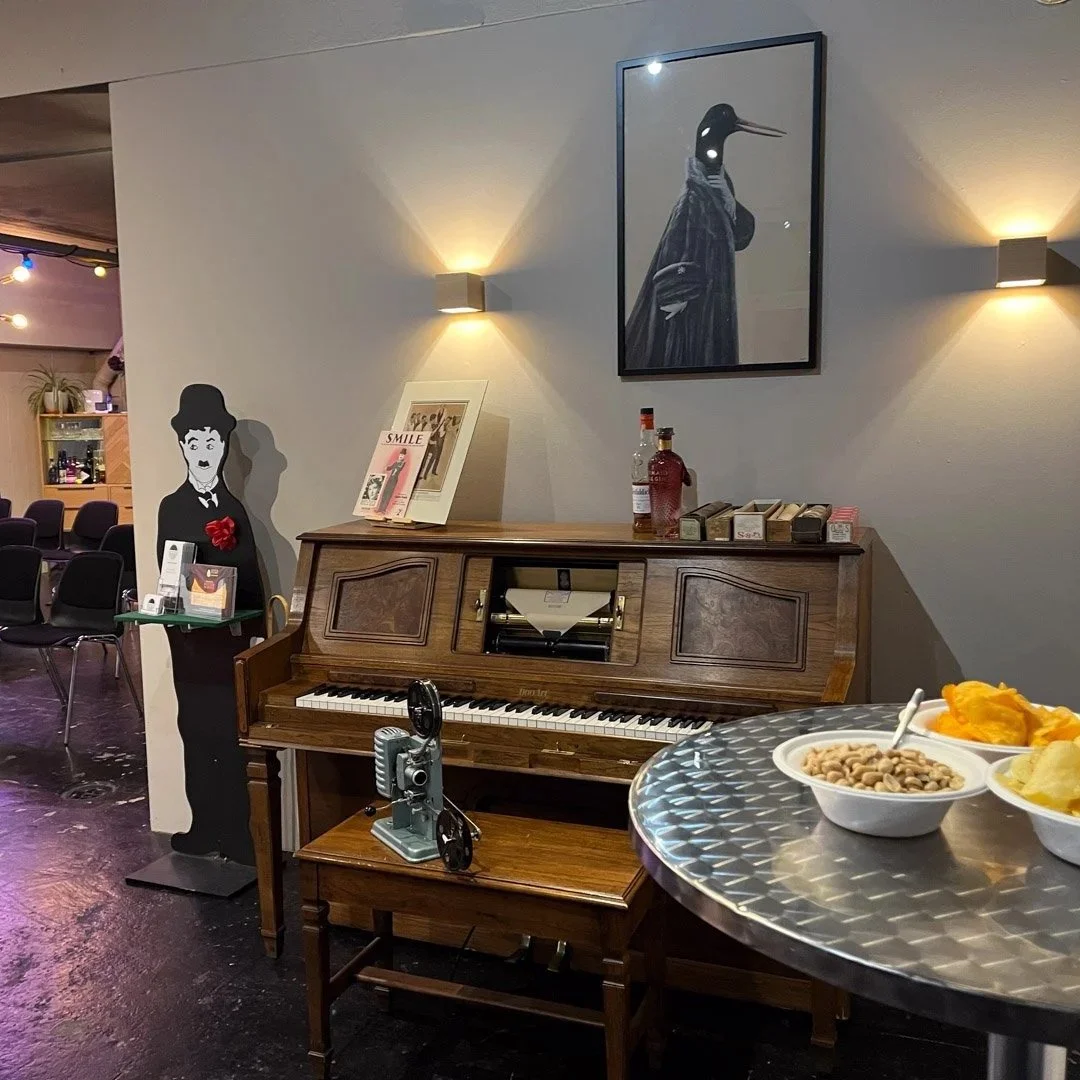 Ein Wohnzimmer mit einem alten Flügel, dekorativem Wandbild und einem stilisierten schwarzen und weißen Mann mit Hut, die mit einem Esstisch voller Snacks im Vordergrund stehen.