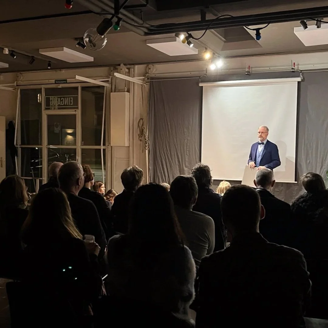 Ein Mann in Anzug und Fliege hält eine Rede vor einem Publikum in einem dunklen Raum vor einer Leinwand.
