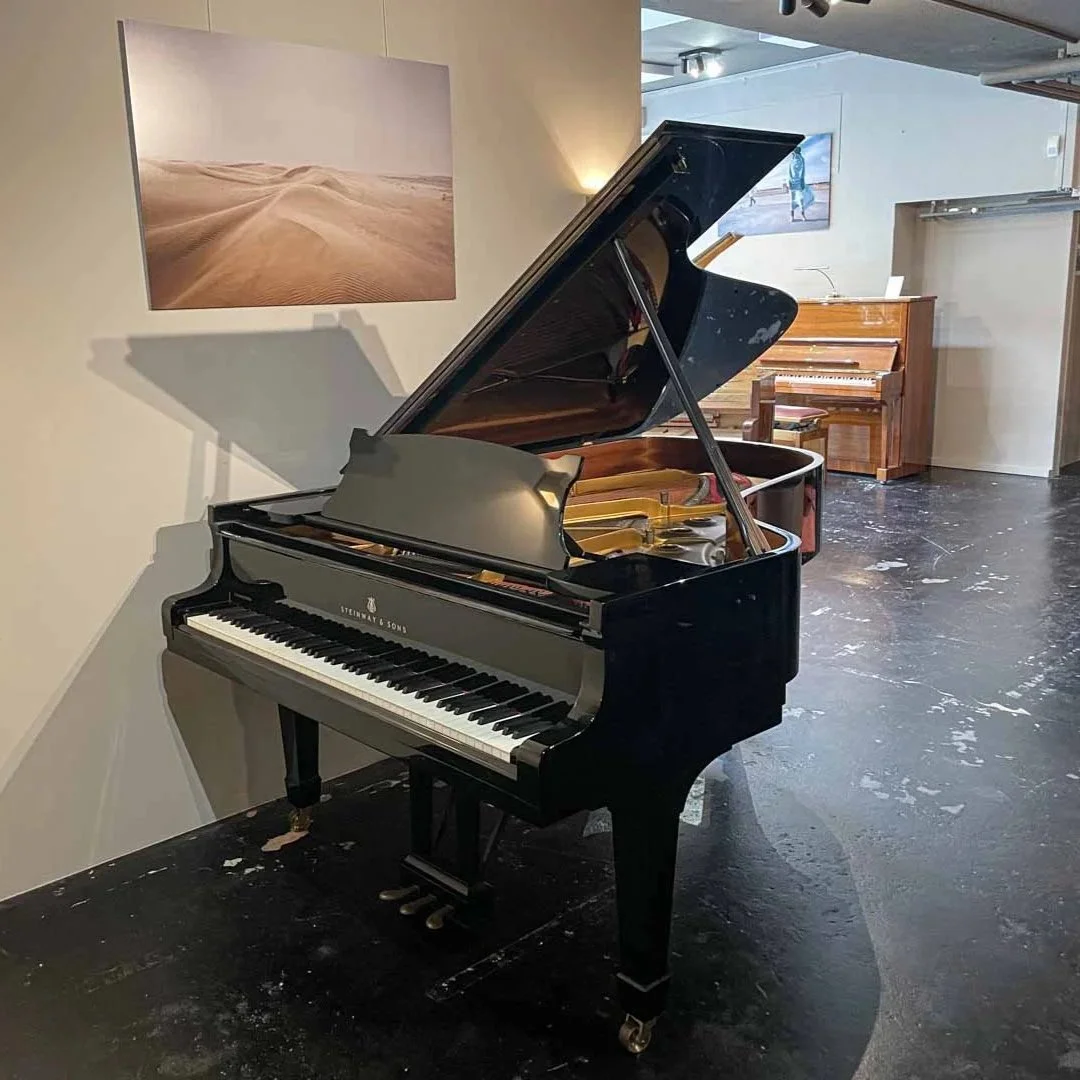 Ein schwarzer Flügel der Marke Steinway & Sons in einem Raum mit dunklem Boden, einem Bild von einer Wüste an der Wand und einem weiteren, kleineren Flügel im Hintergrund.