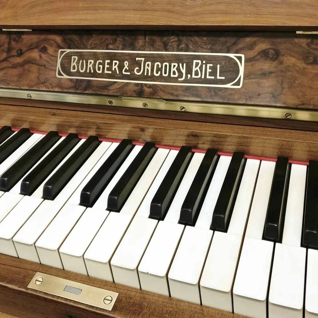Nahaufnahme eines Klaviers mit einem Holztastaturhaube. Auf der Haube steht in weißen Buchstaben "BURGER & JACOBY, BIEL".