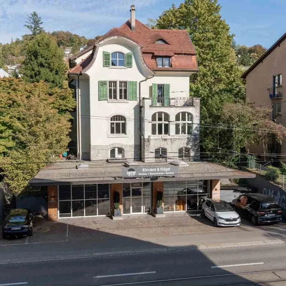 Mehrstöckiges Haus mit weißer Fassade, grünen Fensterläden und Satteldach. Unten befindet sich ein Ladengeschäft mit großen Fenstern und einem Schild, das "Klaviere & Flügel" verkauft. Im Vordergrund ist eine Straße mit parkenden Autos.