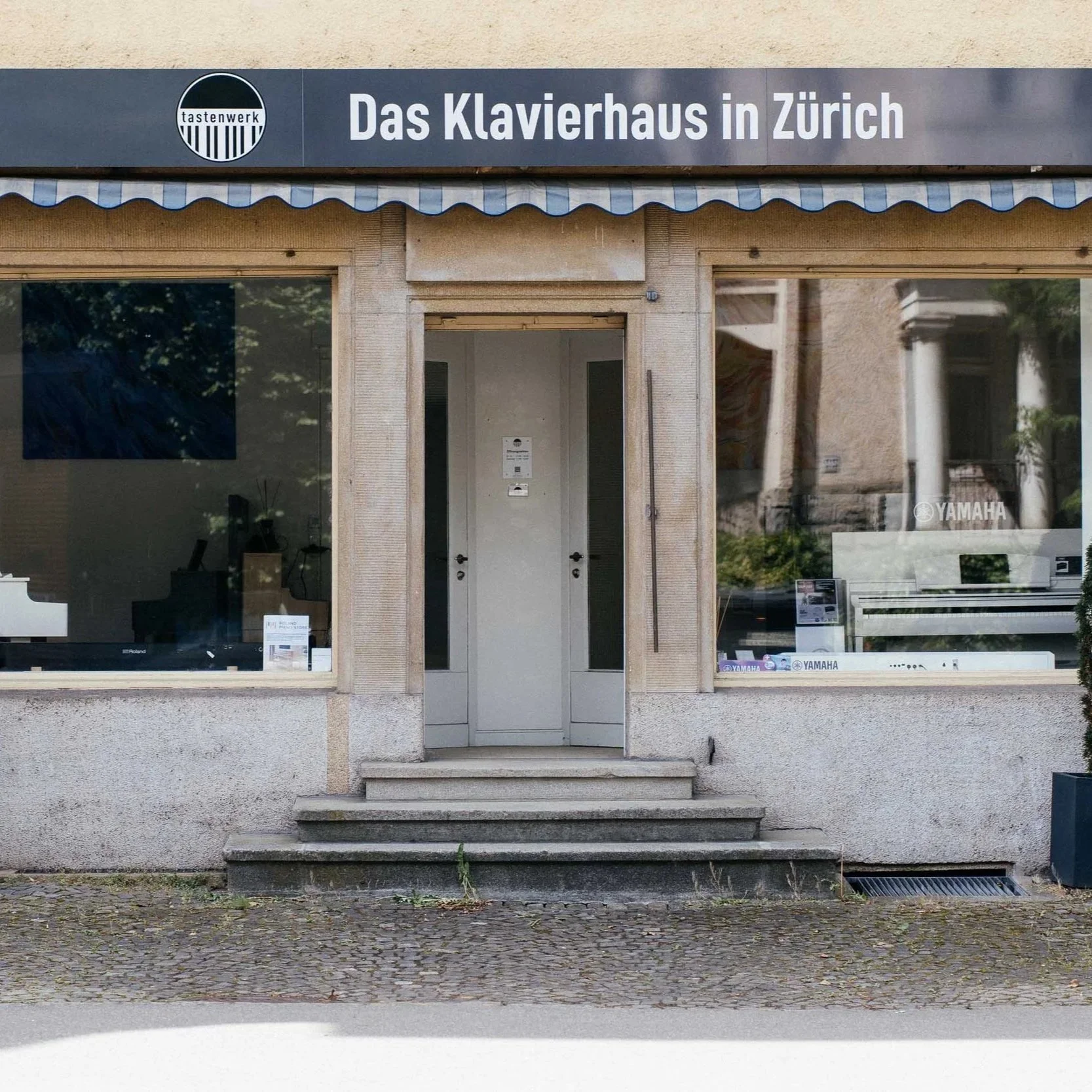 Schaufenster eines Klavierhauses in Zürich mit Eingangstür in der Mitte, zwei große Fenster seitlich, Marken Yamaha und Roland sichtbar, und ein dunkelblauer Schriftzug 'Das Klavierhaus in Zürich' darüber.