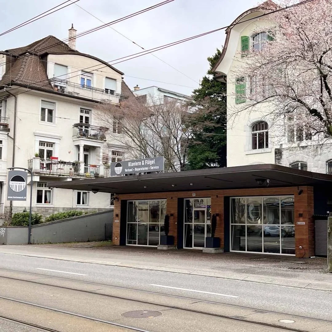 Modernes Gebäude an der Straße mit großen Glasfenstern und einer Schiebetür, im Hintergrund alte weiße Gebäude und blühende Bäume.
